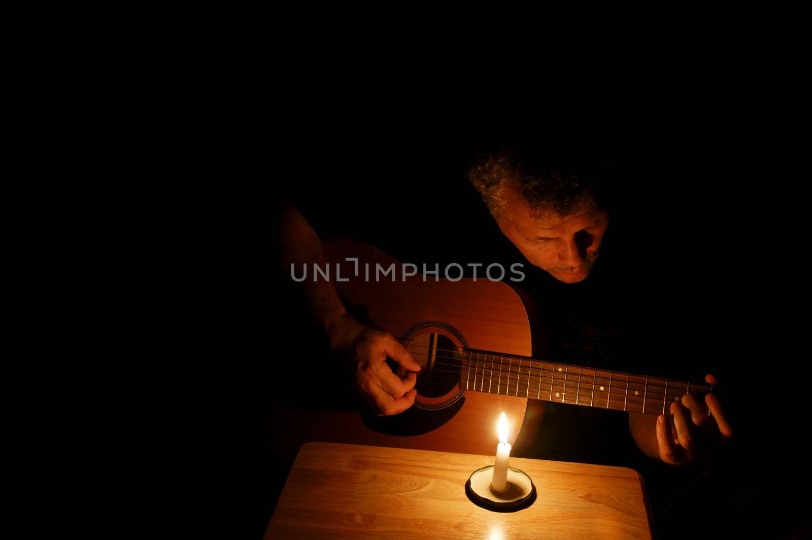 A middle-aged man playing his guitar at night by candlelight.