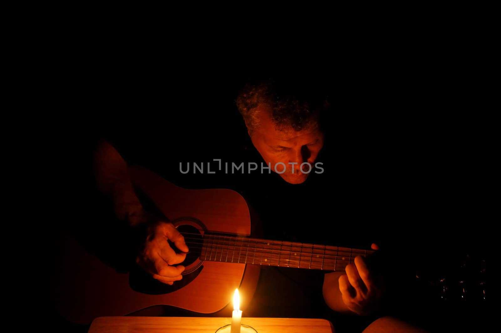 A man playing his guitar at nightime with just the light of a single candle.