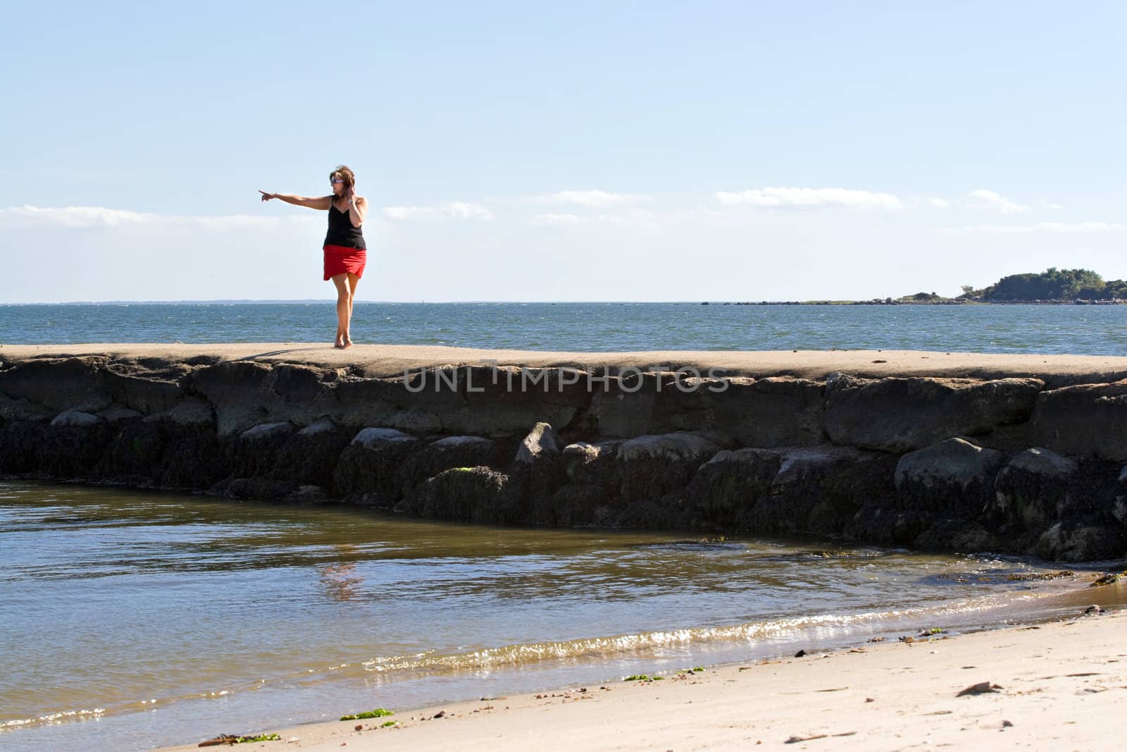 A woman walks down the jetty at the beach pointing to something in the distance.