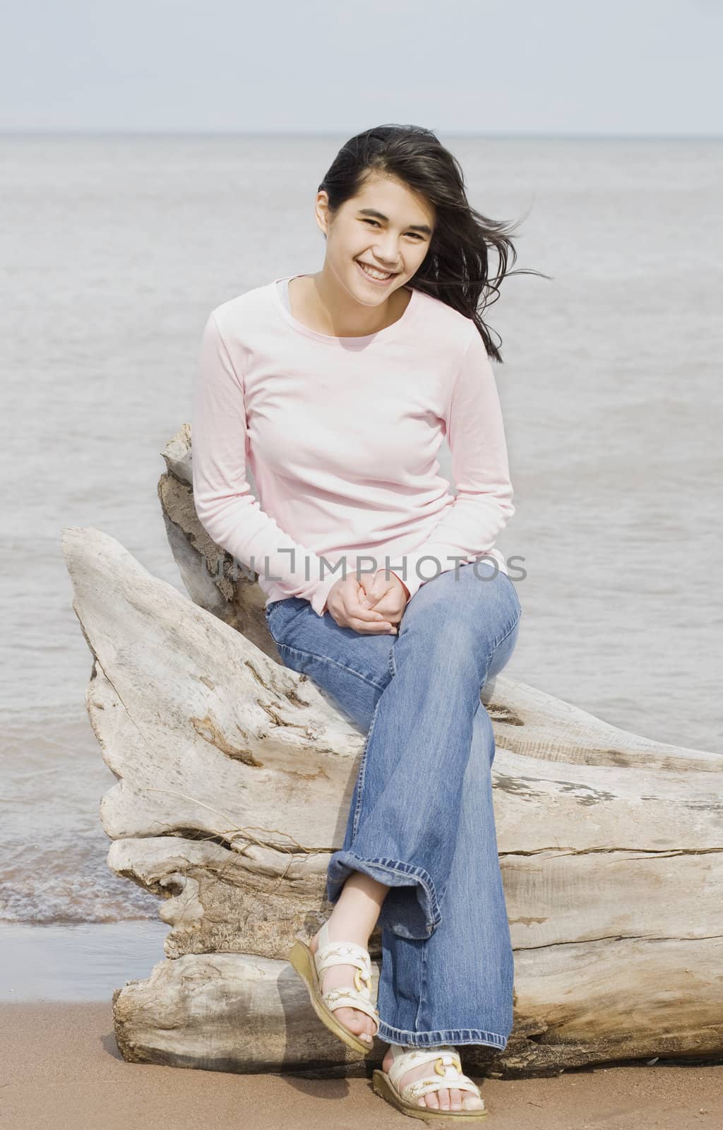 Beautiful biracial teen girl sitting on fallen log by lake shore in summer