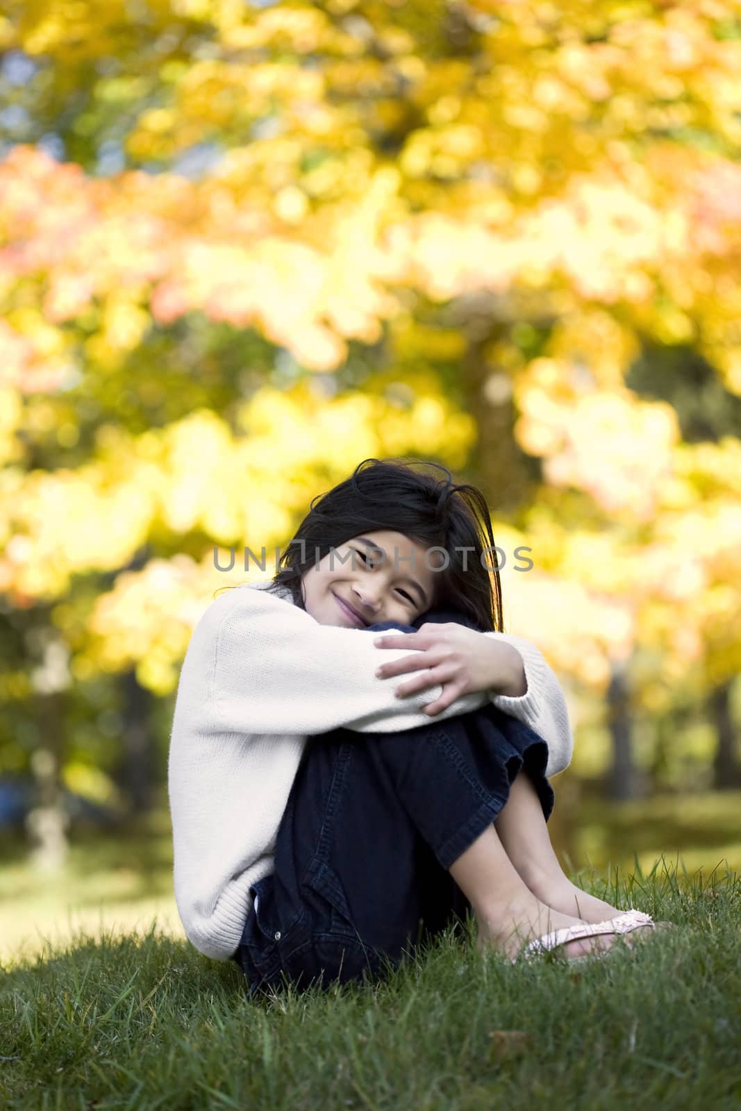 Little girl hugging knees sitting on lawn against bright autumn leaves in background