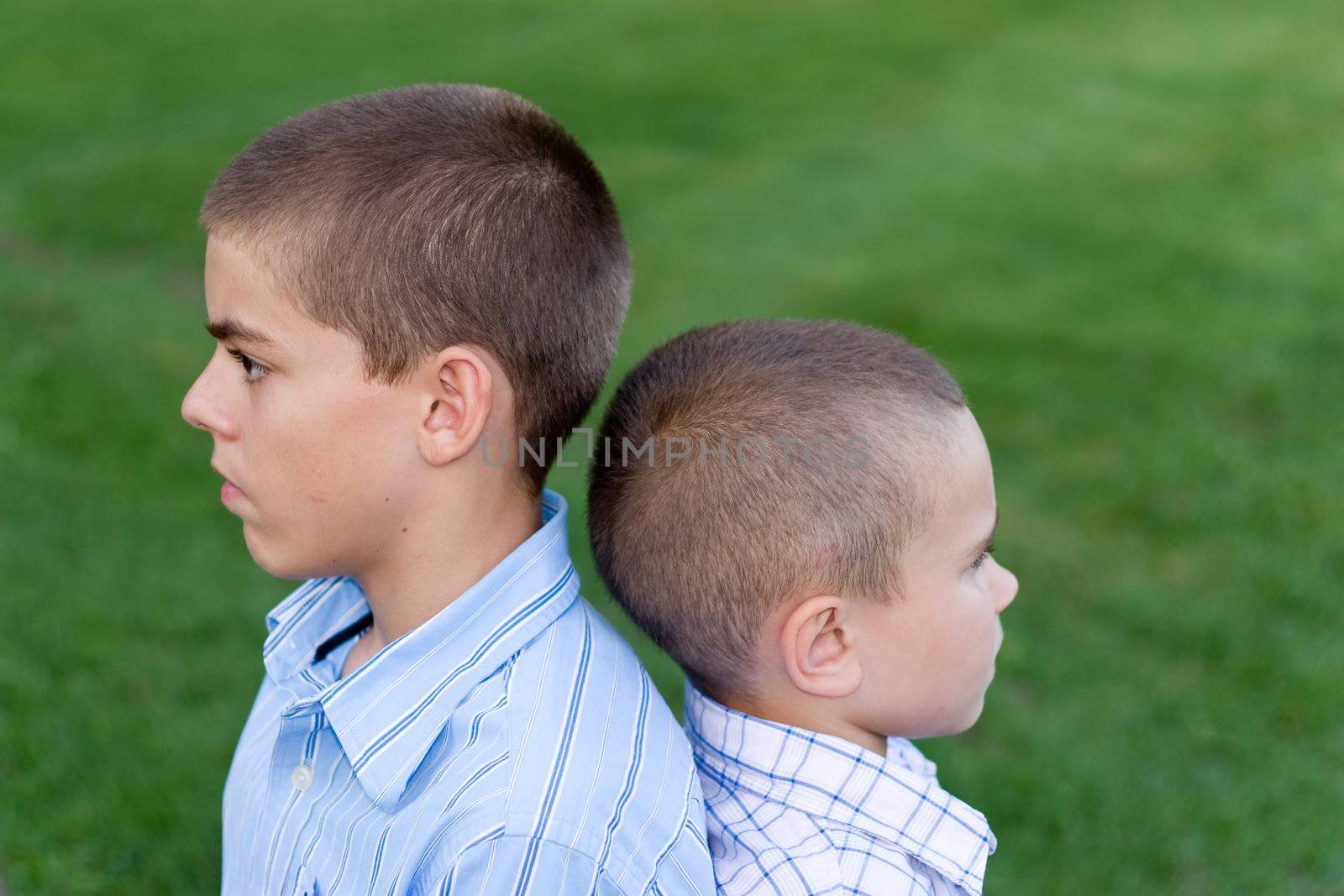 Two young boys sitting back to back outdoors.  