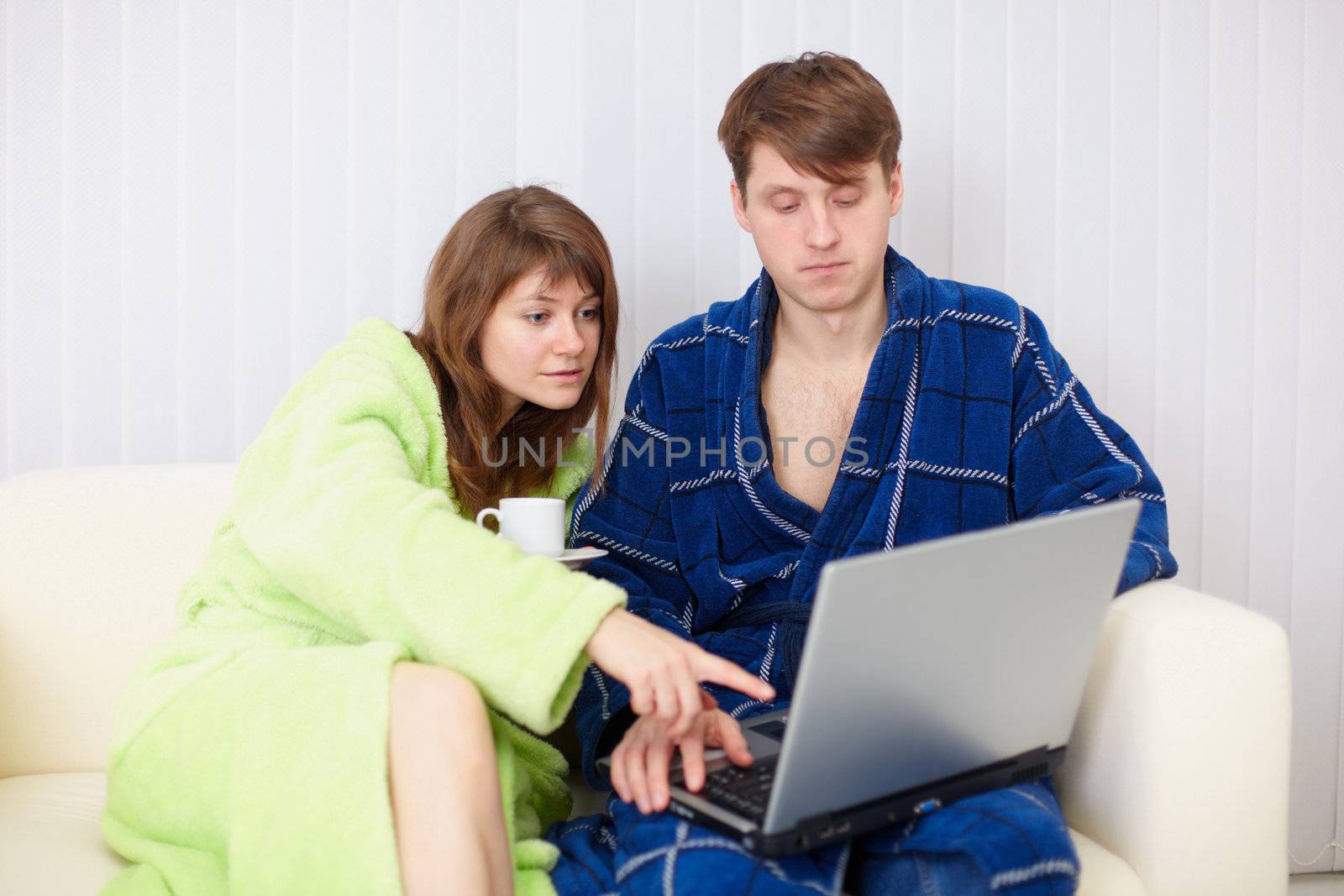 Spouses sit on sofa in dressing gowns with laptop by pzaxe