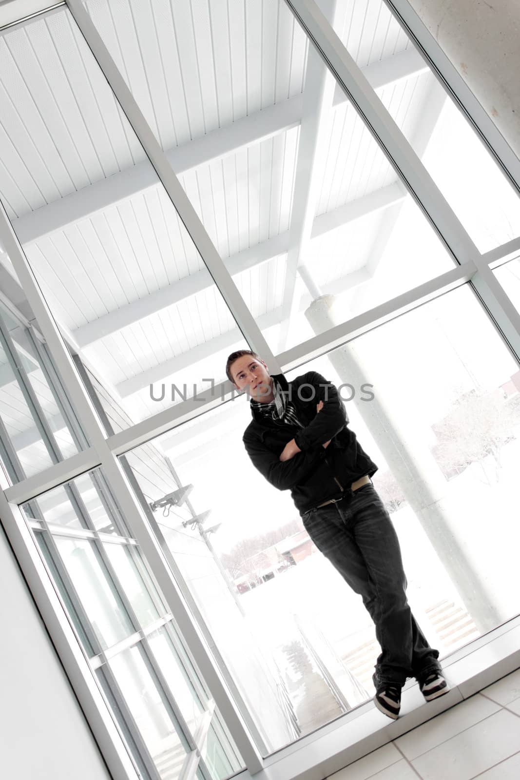 A young man standing by some large windows alone with his arms crossed.