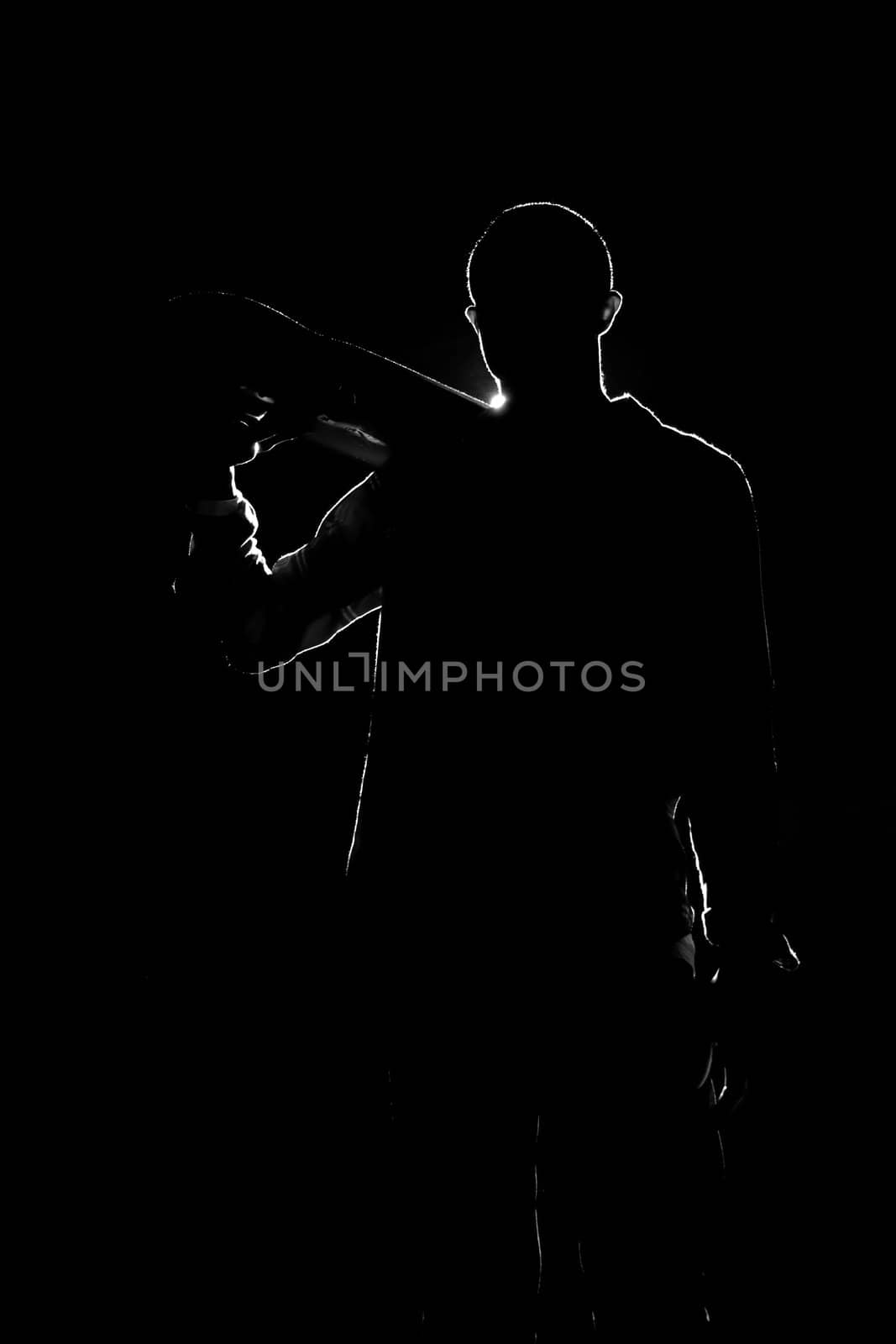 A backlit skateboarder guy posing under dramatic rear rim lighting with his skateboard in black and white.