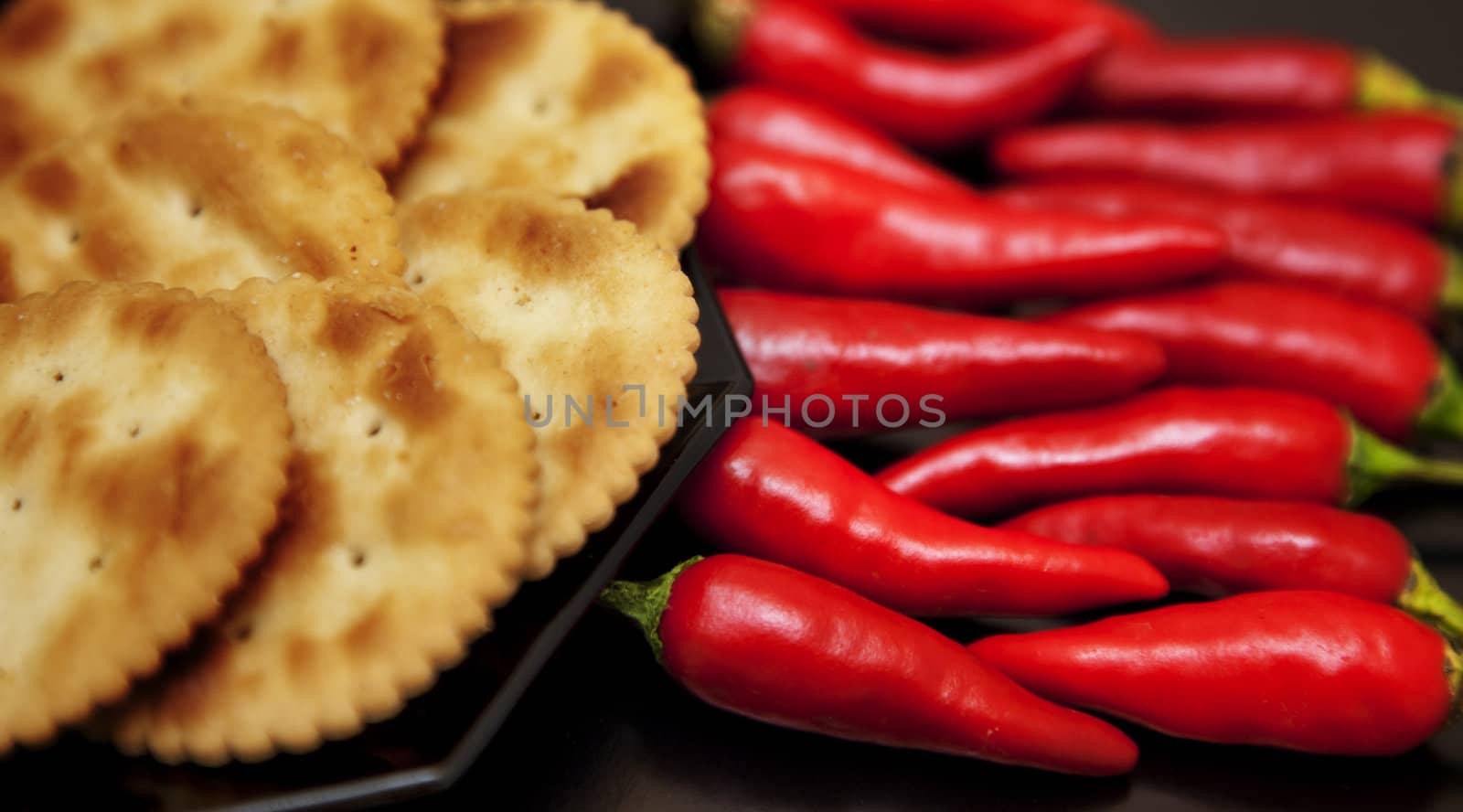 A plate of crackers next to a bunch of fresh chillies against black.