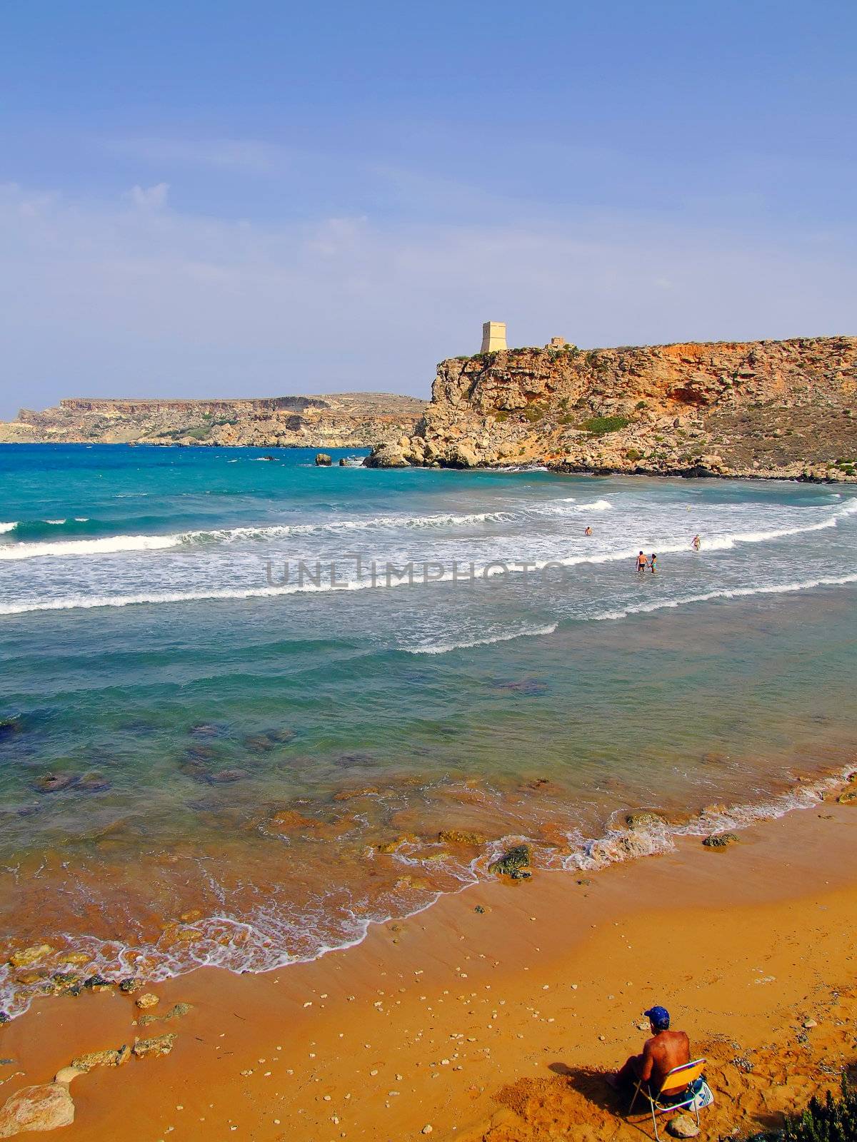 Lone man sitting near the clear blue Mediterranean waters on windy day, with beach and typical rocky coastline