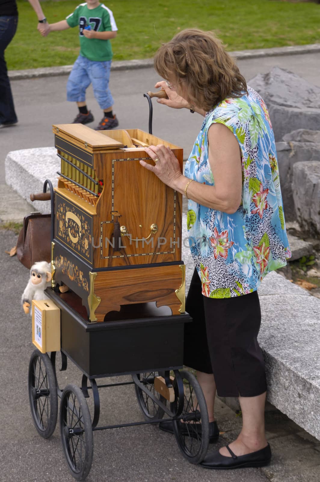 A woman barrel organ player, complete with monkey, playing her music as a man hurries by with his son (who wants to stop and listen).