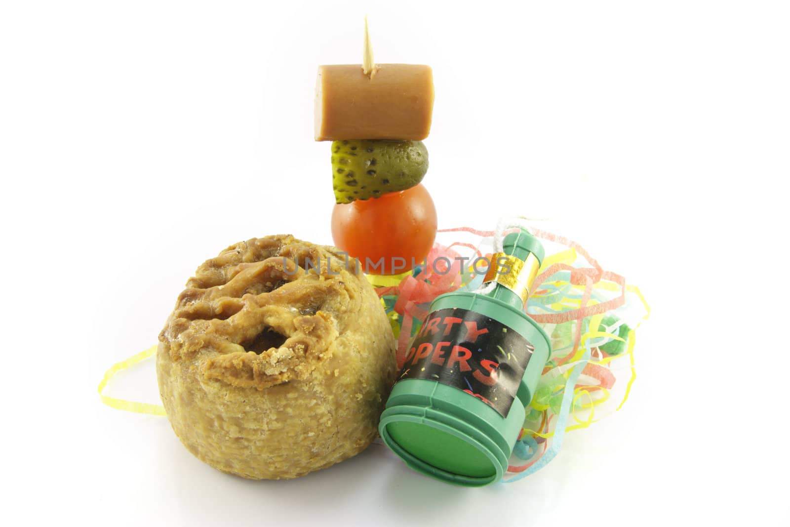 Small pork pie with party popper and cocktail stick containing hot dog sauage, gherkin and tomato with streamers on a reflective white background