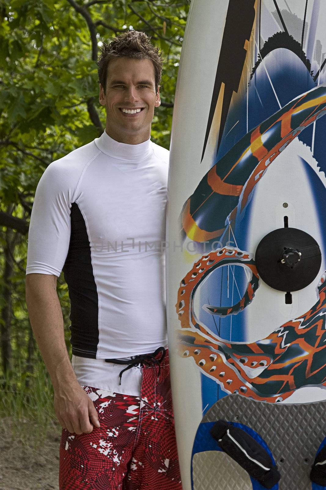 Windsufer standing beside his board in front of trees