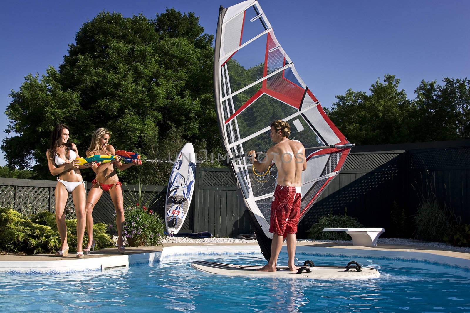 two girl spray a windsufer, in a pool, with water gun