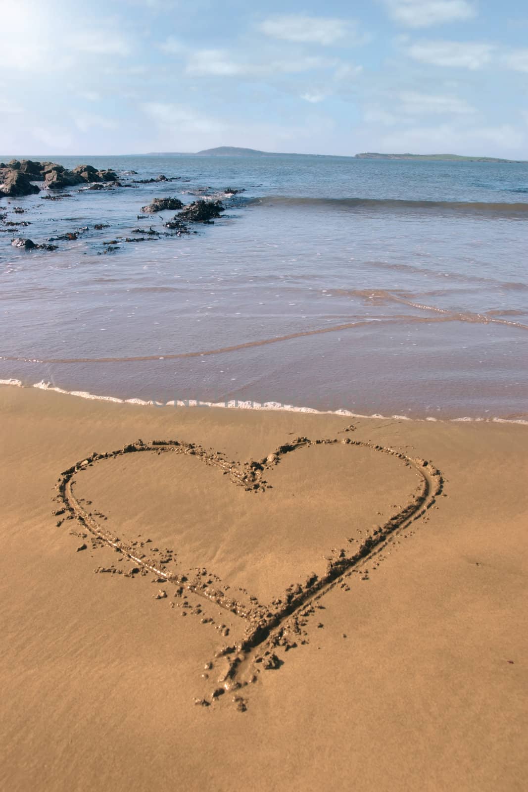 a romantic love heart inscribed on the beach with waves in the background on a hot sunny day