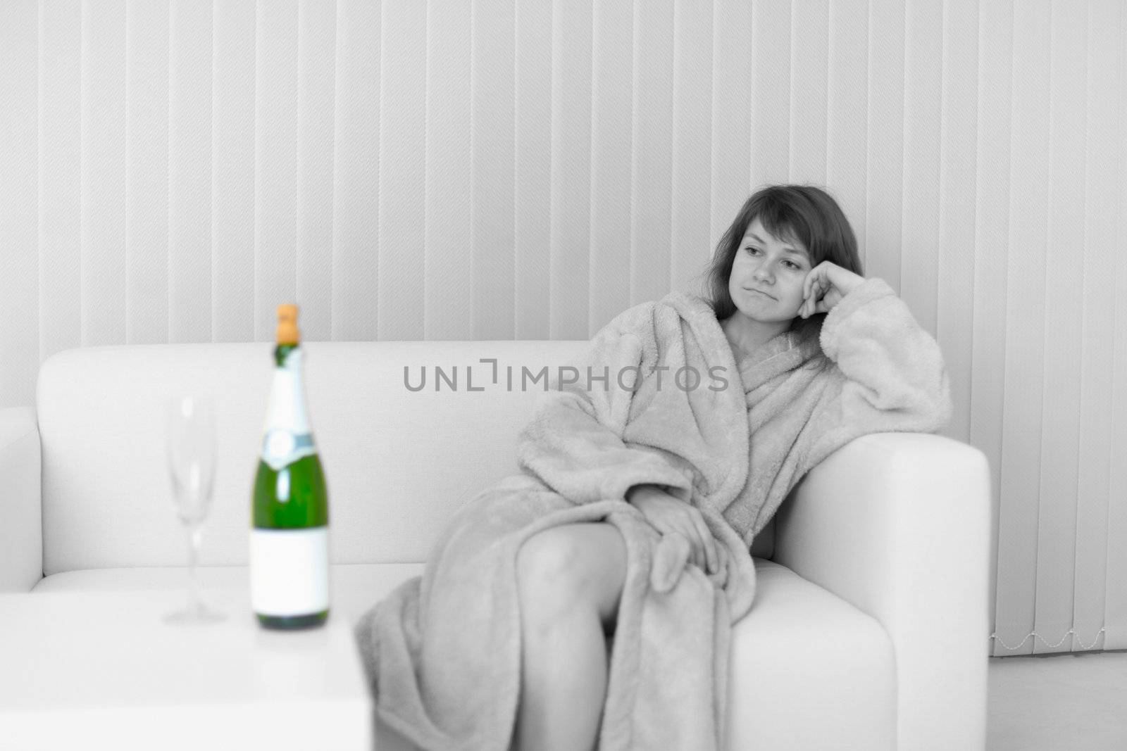 The lonely young woman sits on a sofa and looks at a wine bottle