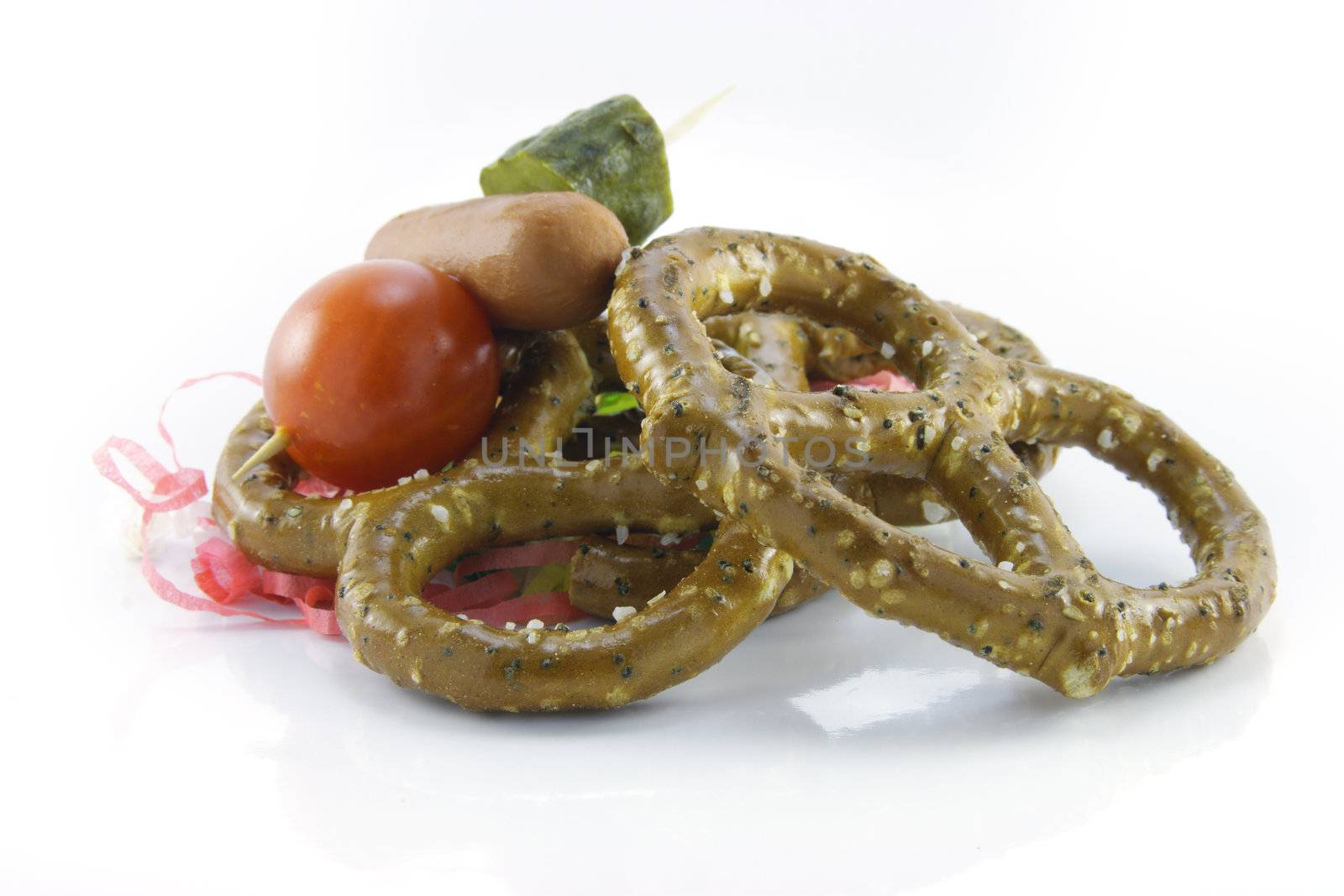 Pile of salty baked pretzels with cocktail stick containing hot dog, tomato and gherkin with streamers on a reflective white background