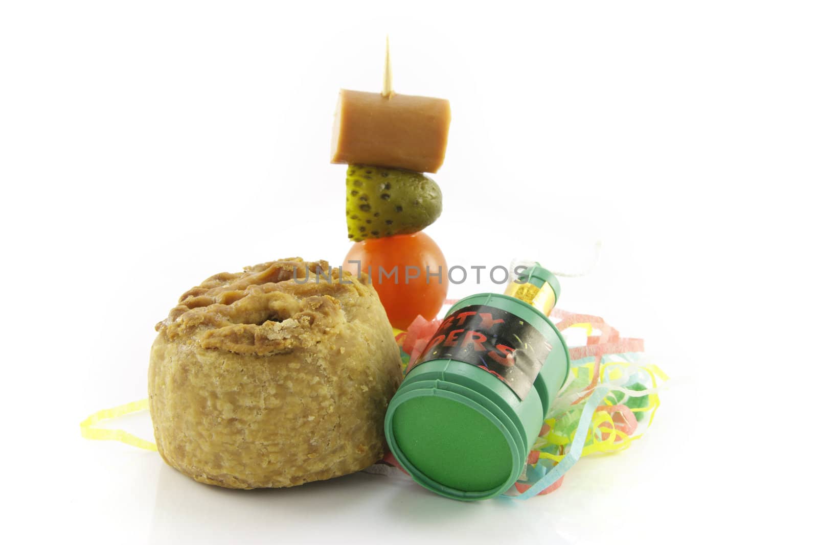 Small pork pie with party popper and cocktail stick containing hot dog sauage, gherkin and tomato with streamers on a reflective white background