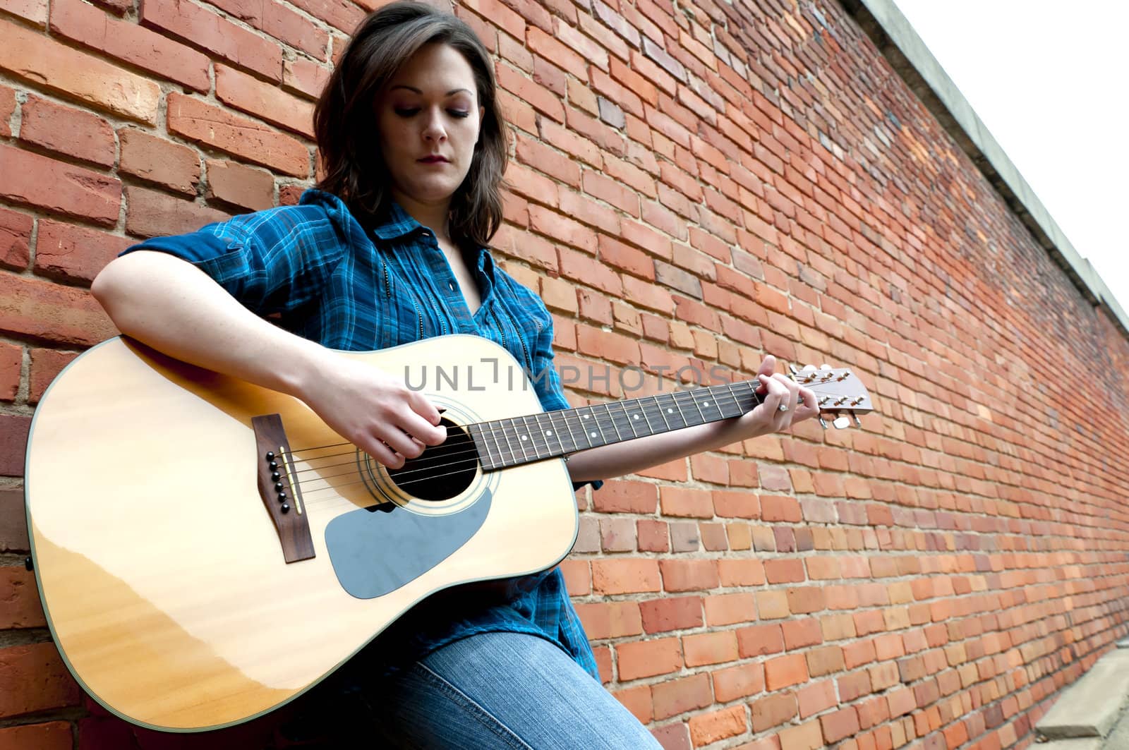 Young woman playing guitar outside while leaning against brick wall.