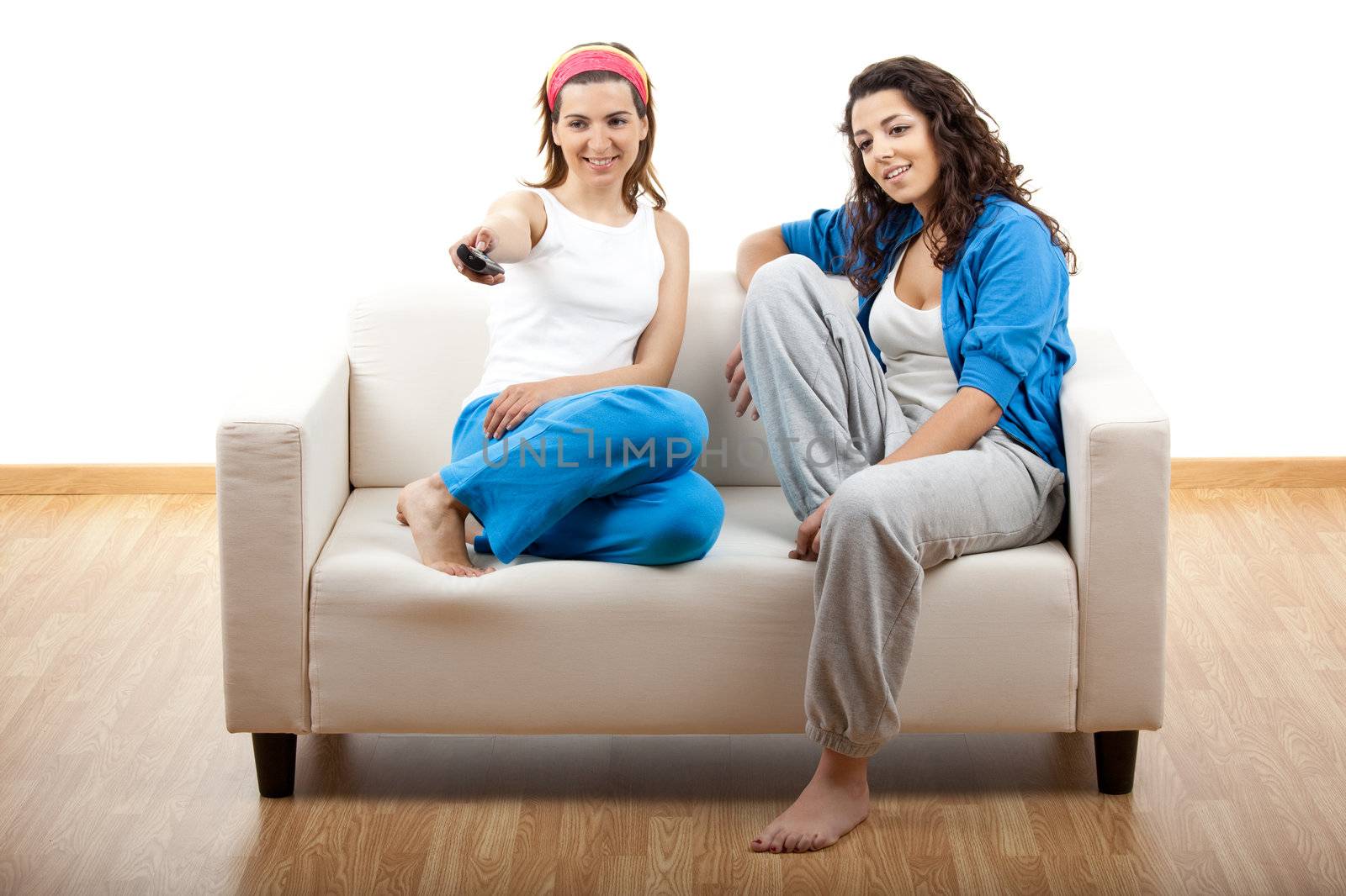 Two beautiful young women relaxing on the couch holding a remote control - isolated on white