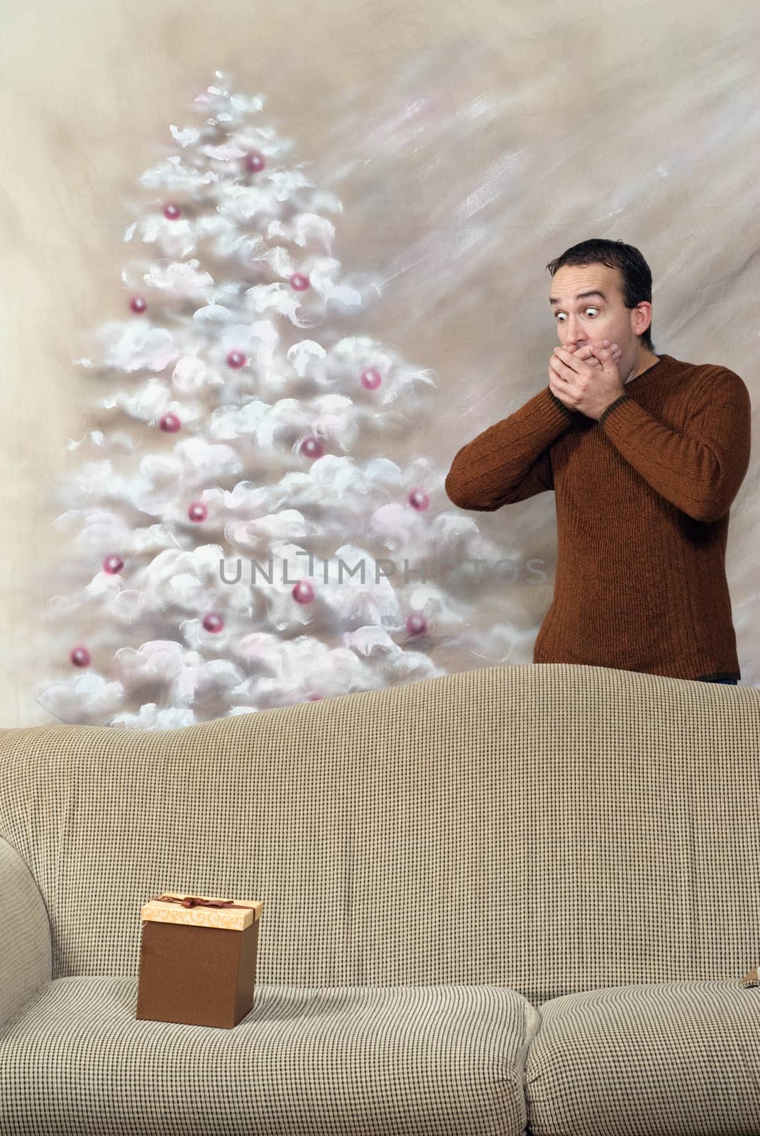 A young man is covering his mouth is shock and surprise that there is a gift on the sofa for him