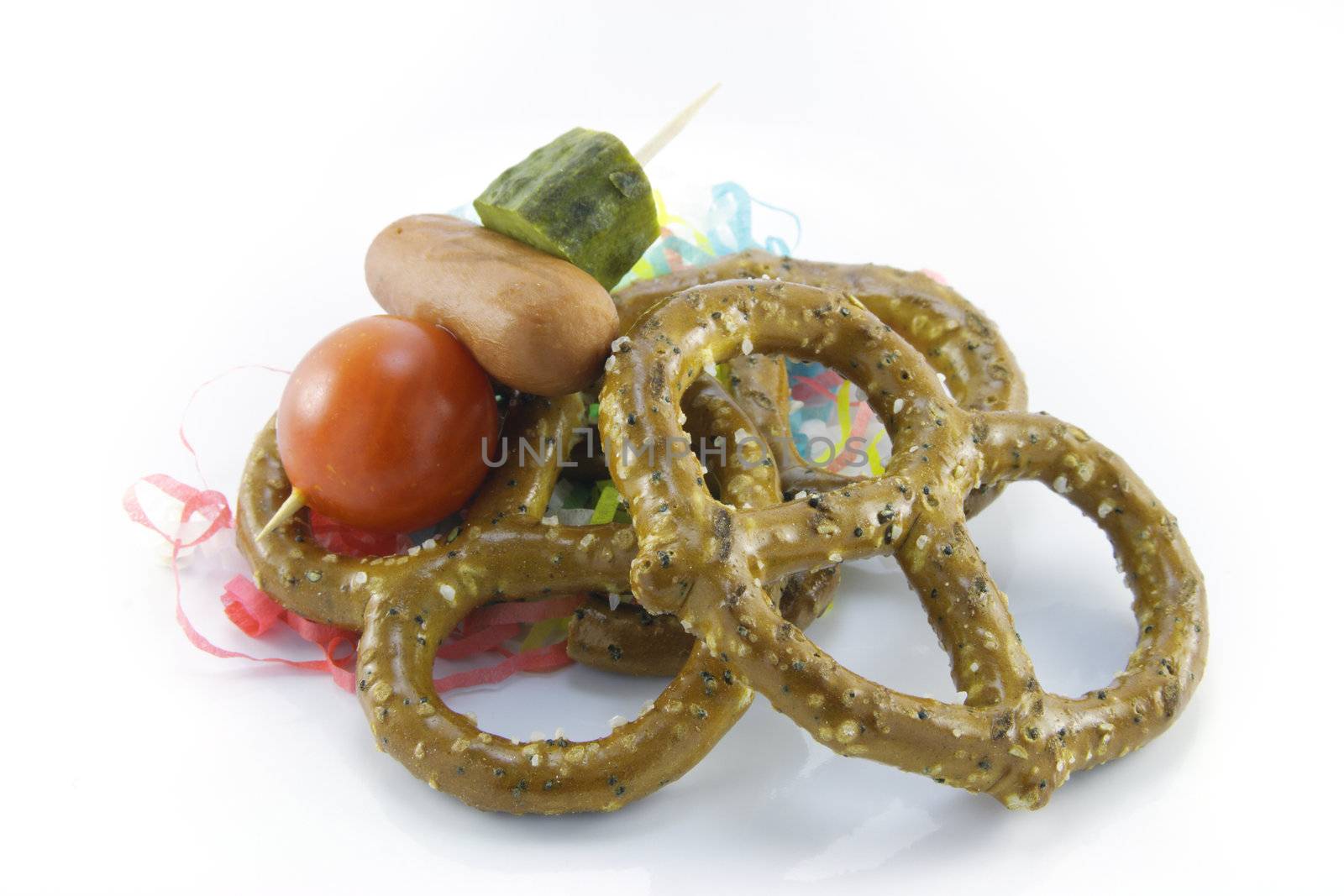Pile of salty baked pretzels with cocktail stick containing hot dog, tomato and gherkin with streamers on a reflective white background