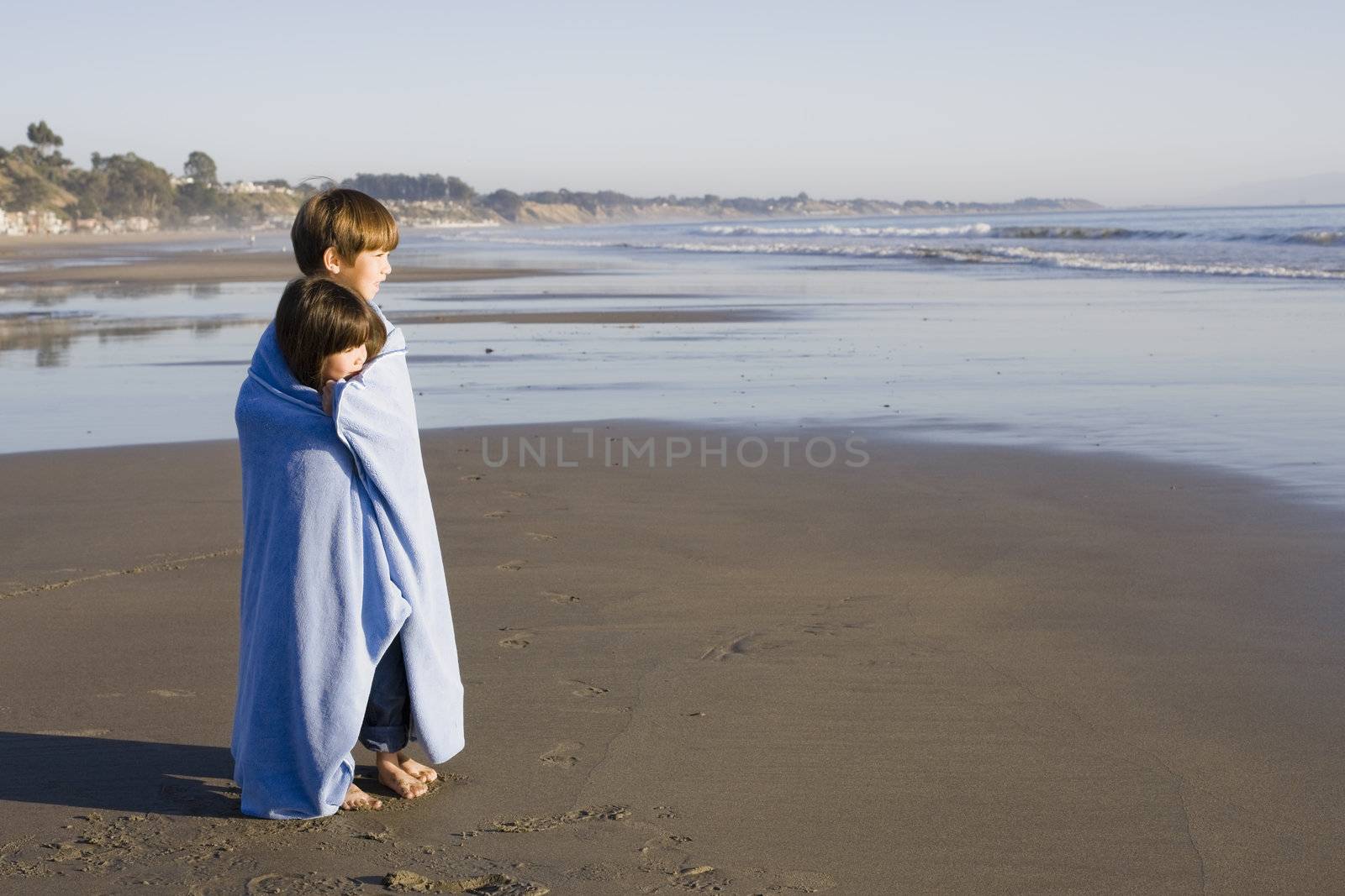 Two Little Kids Standing at Beach Wrapped in a Blanket Looking out to Sea