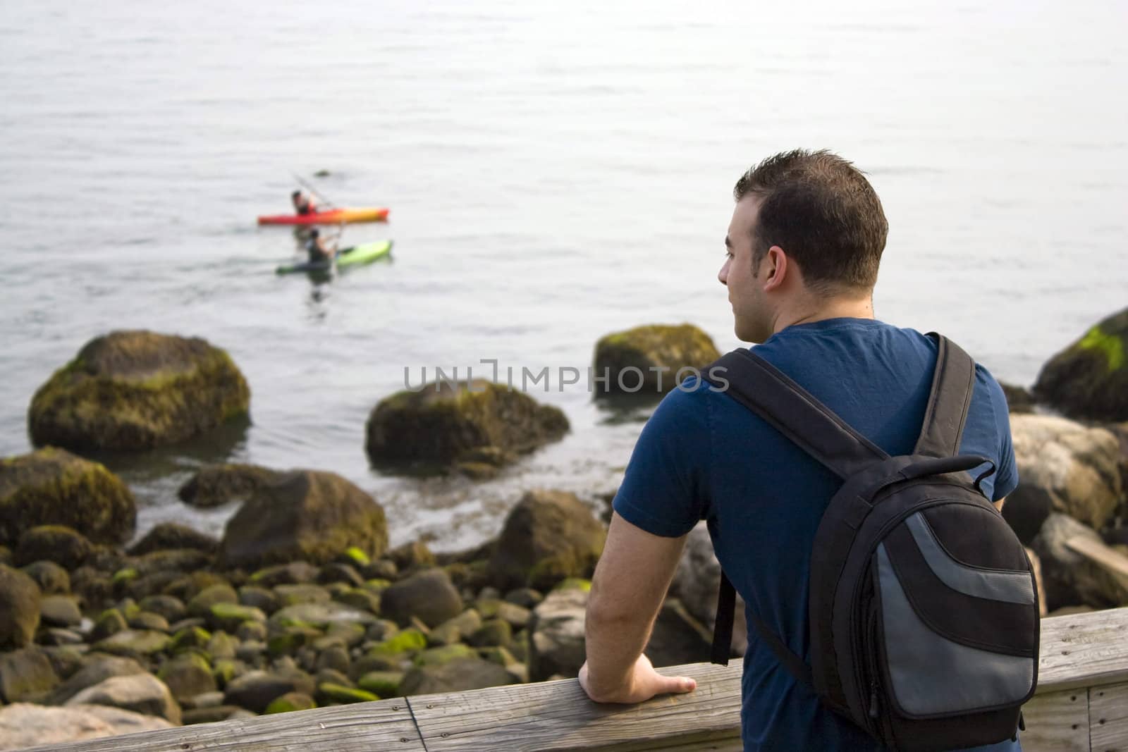 A young man enjoys the view by the sea as two people kayak in the distance.  Shallow depth of field.