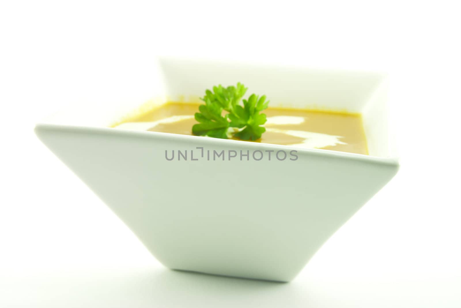 Rich red hot tomato soup in a small square bowl with a sprig of parsley and cream on a white background