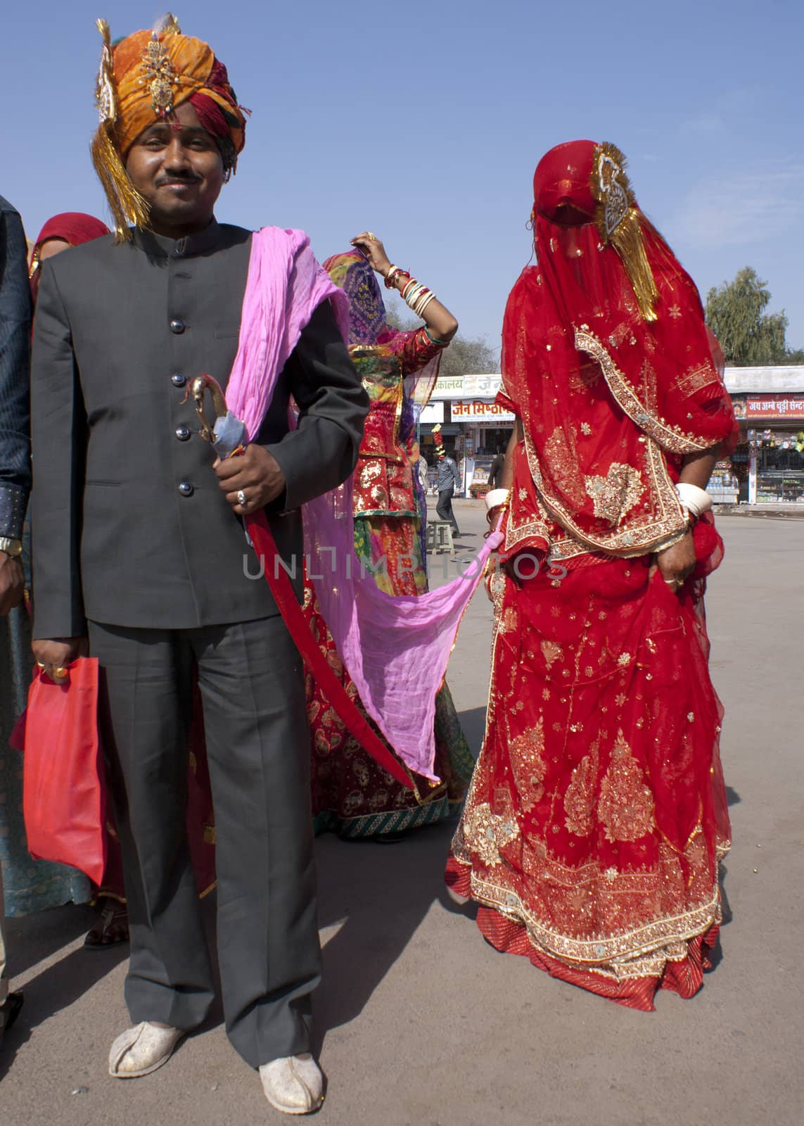 A young fancy dressed boy-man, with a moustache, pulls a totally covered and colorful veiled young woman on a pink leash behind him, while other women and men sing and clap their hands. With his orange-golden turban, overly decorated with precious stones, and his Mao-style dark grey suit closed up to his chin, over white shoes, he radiates triumph and smiles happiness for all to see.