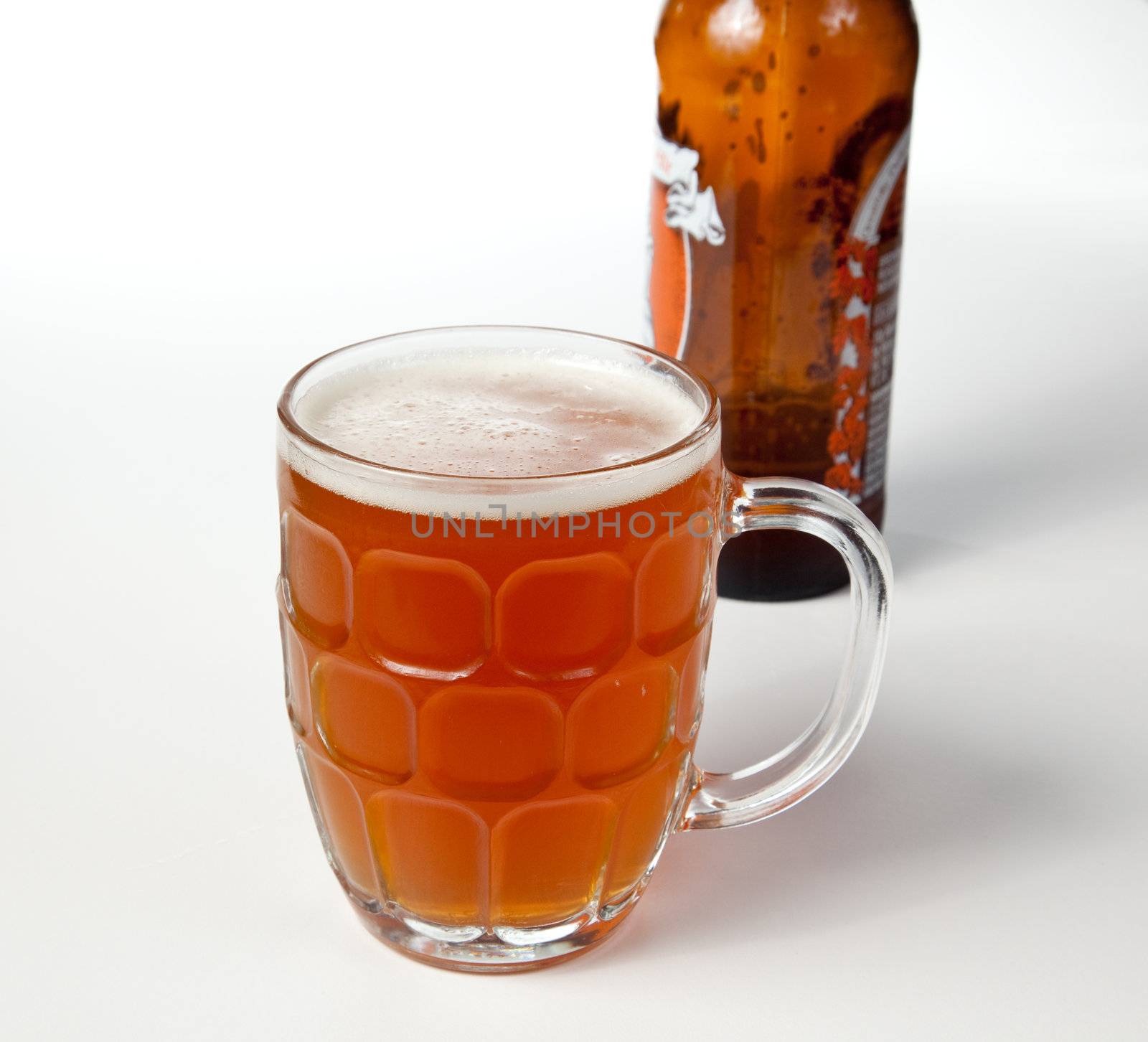 Golden brown beer in an English style pint mug with foamy head and bottle out of focus in background