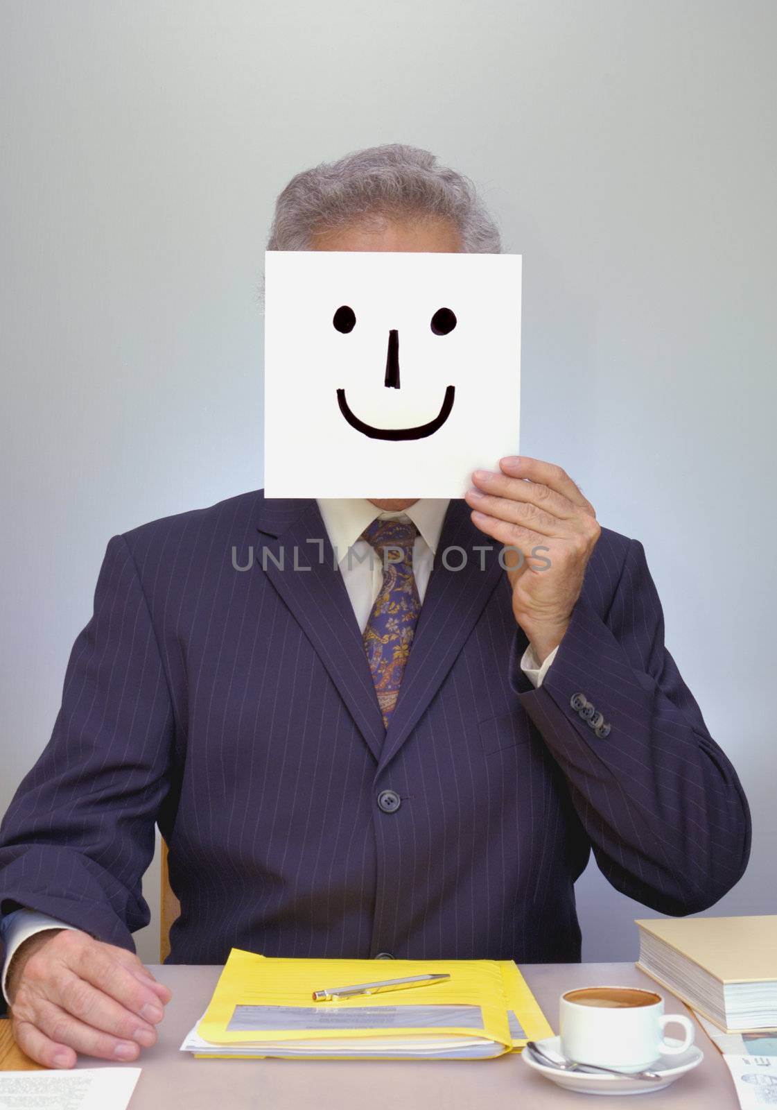 A businessman sits at his desk, holding a home-made smiley mask in front of his face. Look ... I'm happy ... really!