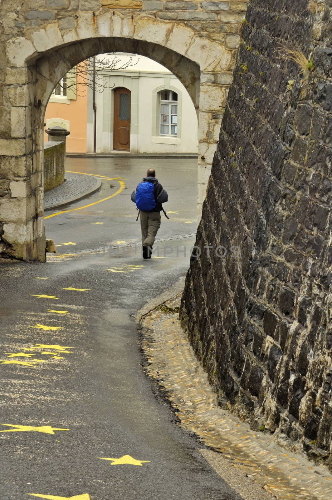 A hiker walks under the arch in the Swiss village of Aubonne on a wet day. Slight motion blur on his feet and backpack straps.