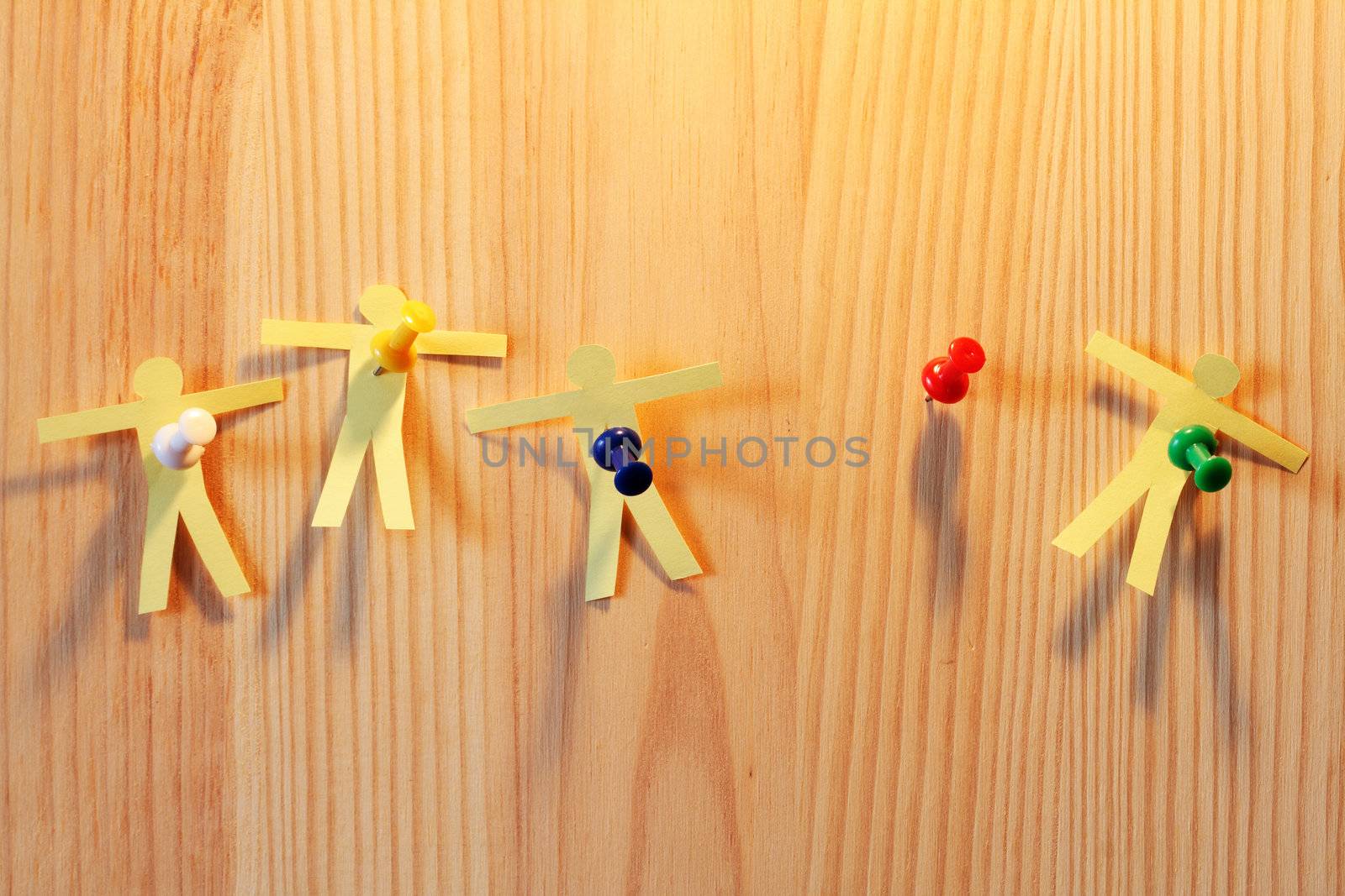 Four paper men hanging with thumbtacks on wooden background. One thumbtack is free