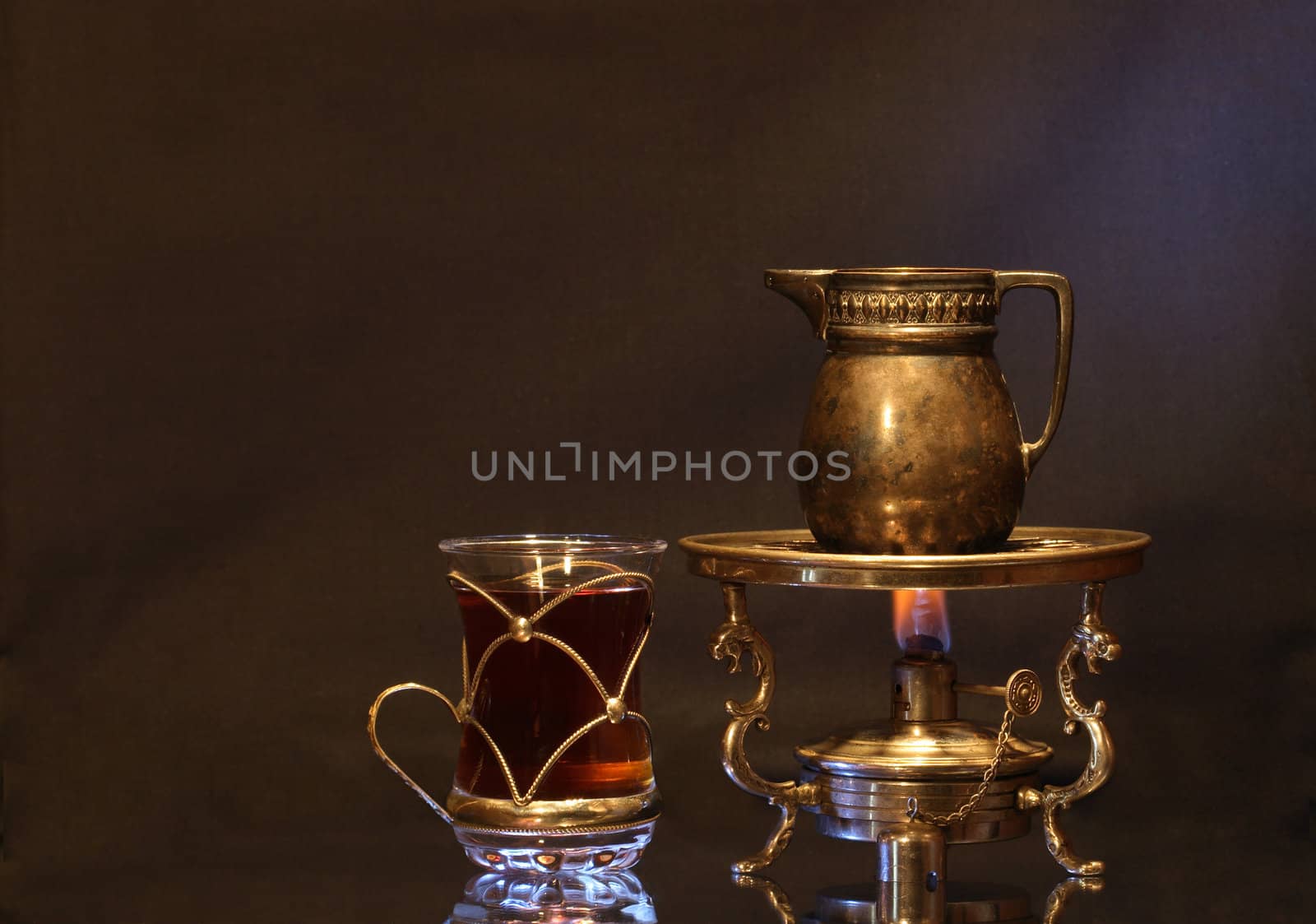 Cup of tea near ancient brass jug standing on vintage table spirit lamp with flame