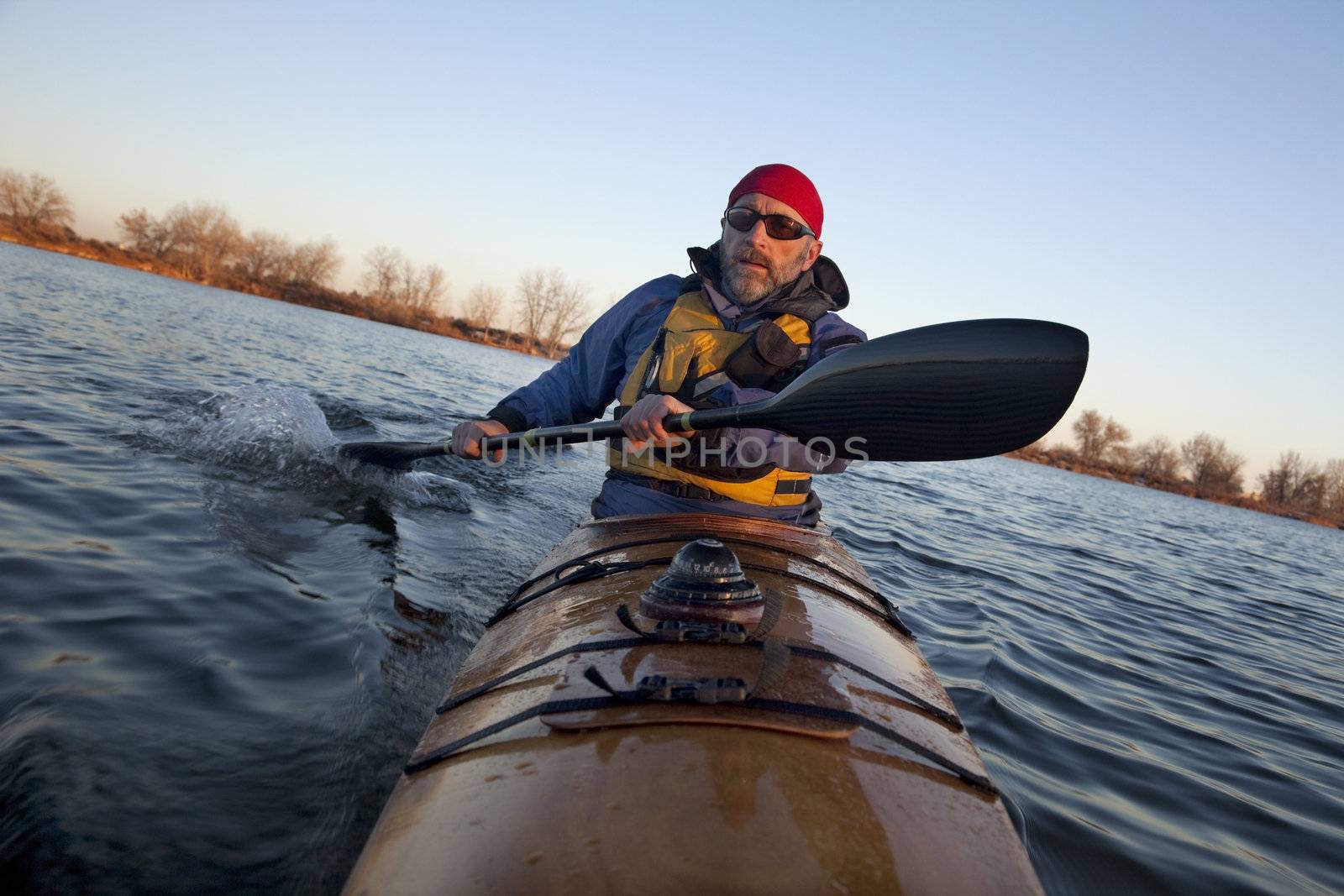 mature male paddler exercising (turning boat using rudder stroke with his wing carbon fiber paddle) in a home built wooden sea kayak on lake, fall scenery  in Colorado, view from kayak bow