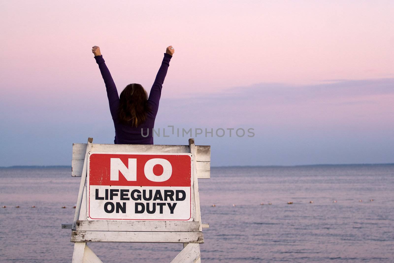 A woman holds her arms up in the air while seated in a vacant lifeguard chair at the beach.