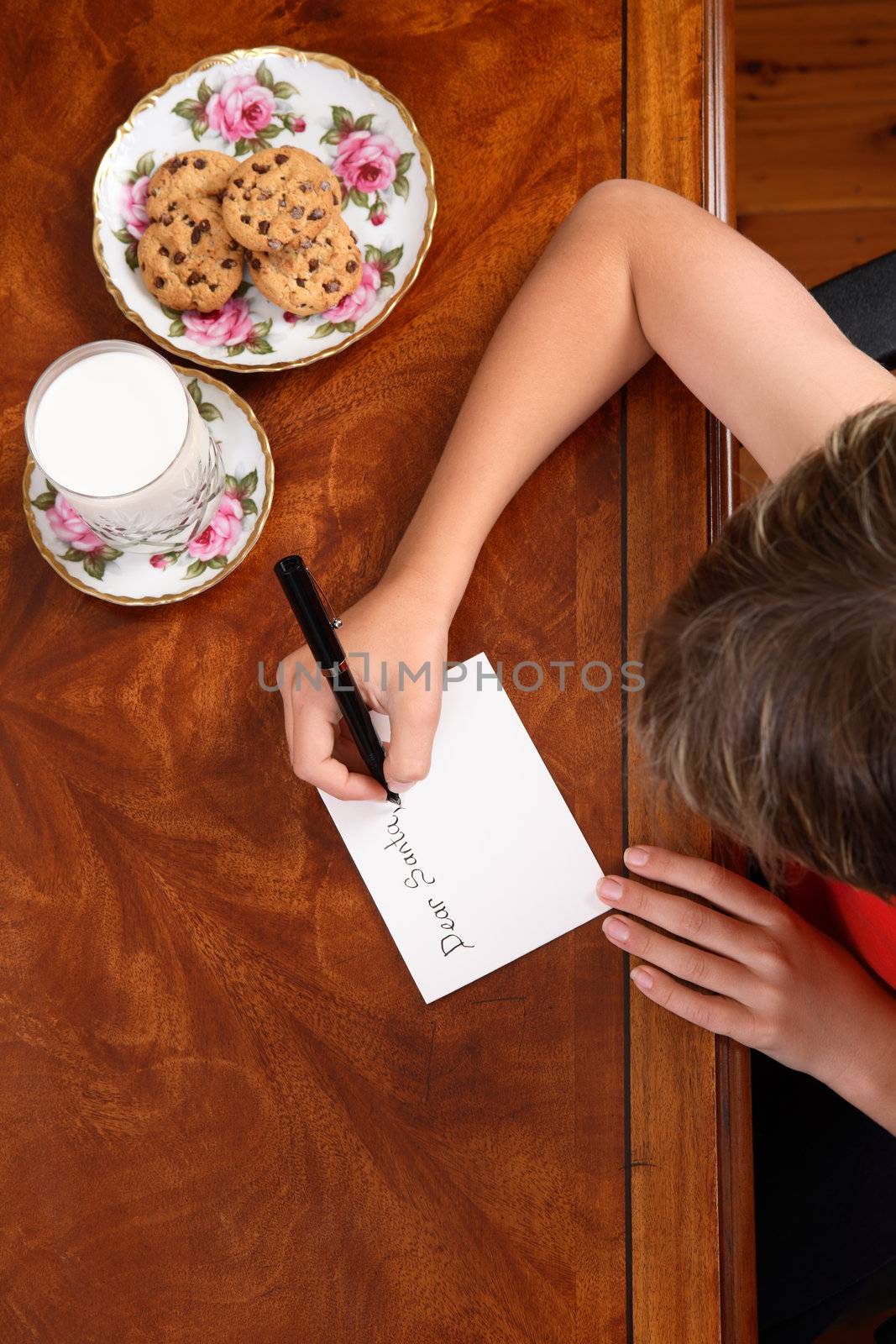A child at desk writing a letter to Santa Claus, beside him a plate of choc chip cookies and glass of milk.