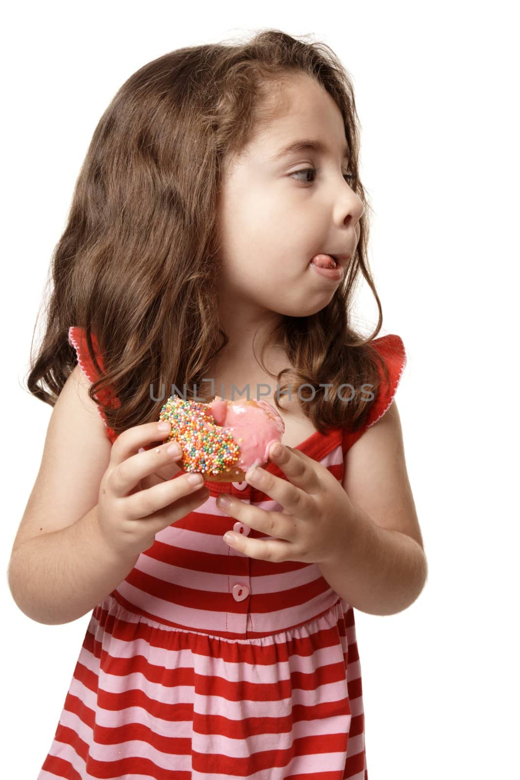 A little girl enjoys a sweet pink iced doughnut.   She is licking her lips and looking sideways.  Suitable for copy.