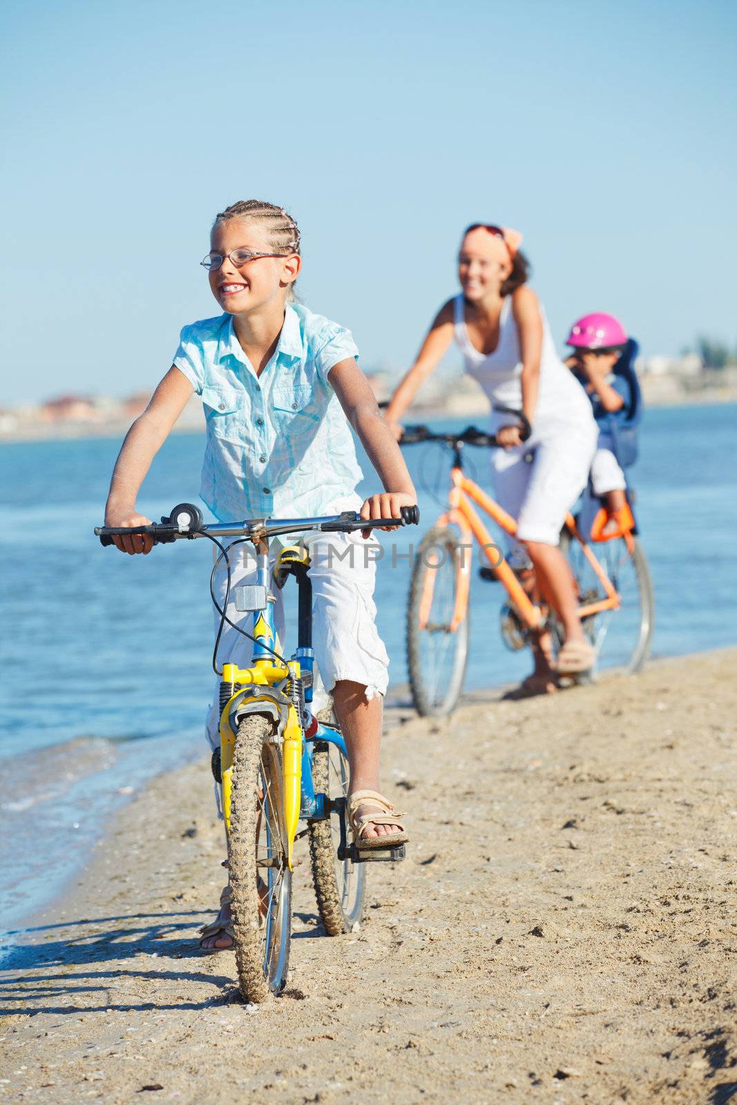 Cute girl with her mother and brother ride bikes along the beach. Focus on girl. Vertical view