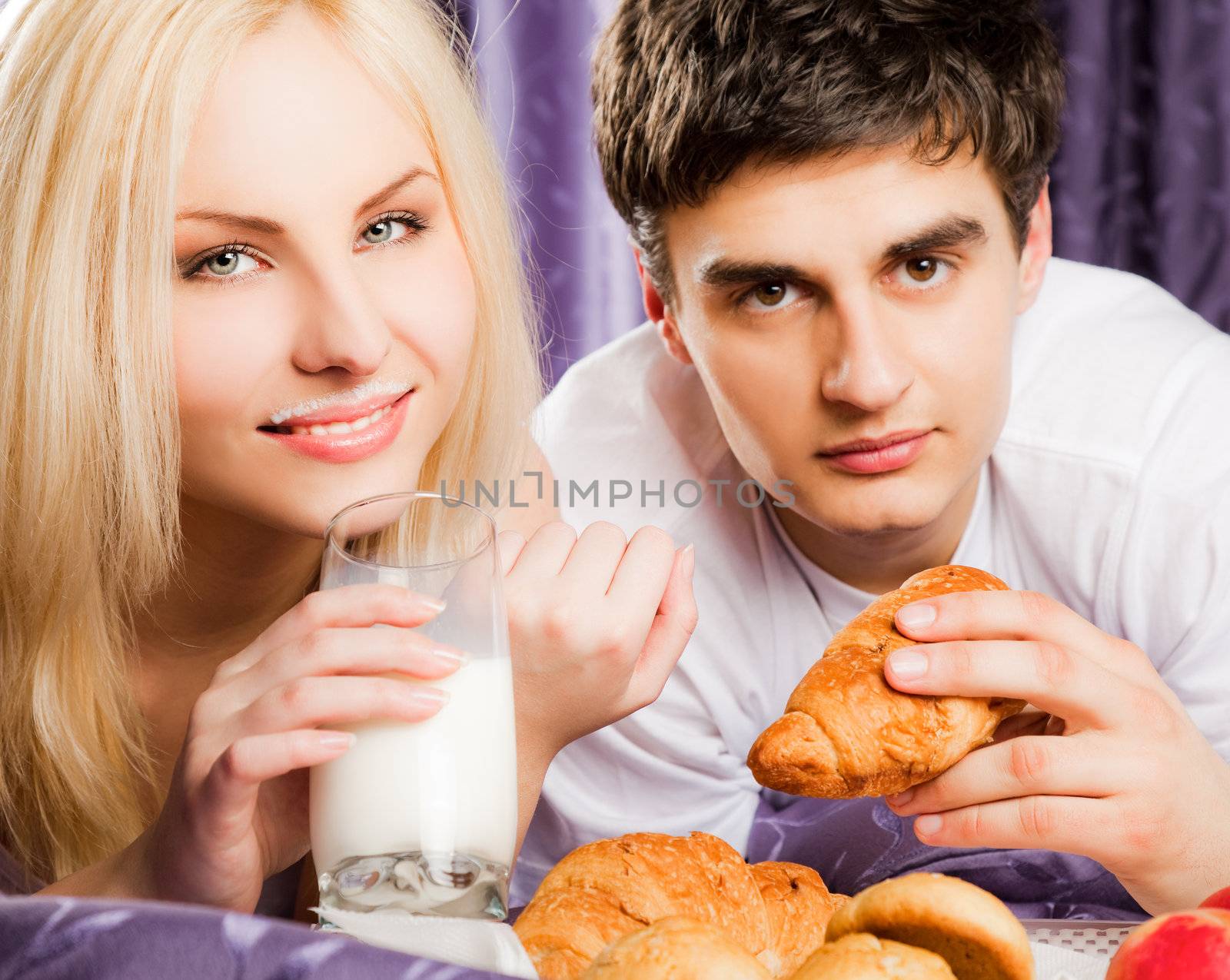 Young couple having breakfast in bed - man eating croissant, female with milk moustache