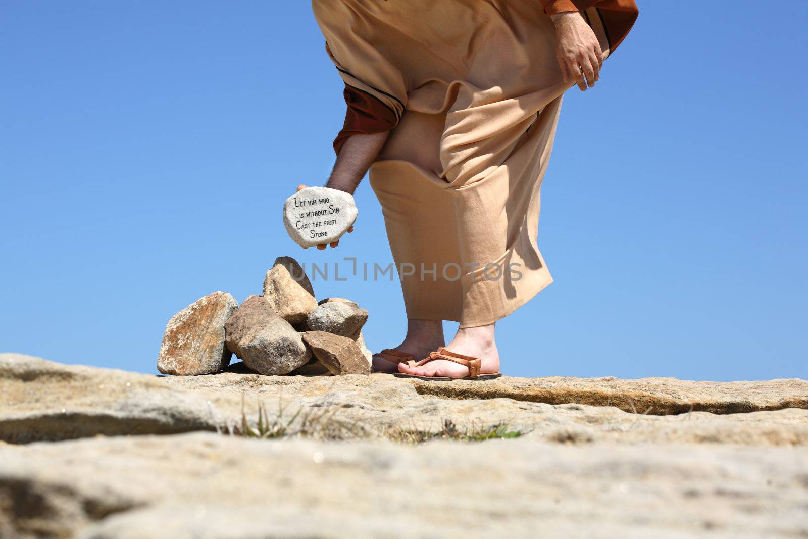 Man standing in the sun on sandy rocks holding a rock depicting ansswer by Jesus  from John Chapter 8. The pharsees were trying to enforce Leviticus 20: 10 however they overlooked Deuteronomy 1: 16, 17. They were more concerned with entrapping Jesus.   Concept of sin, mercy, forgiveness, love, conscience, human rights, 