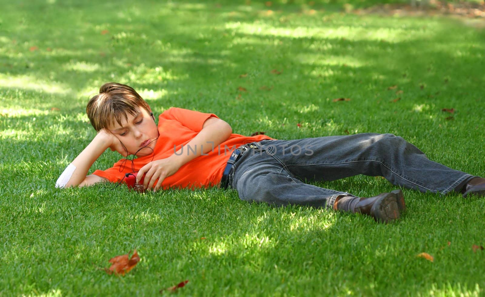 a young boy sprawled out on the shaded grass in the park, listens to music on an mp3 music player.