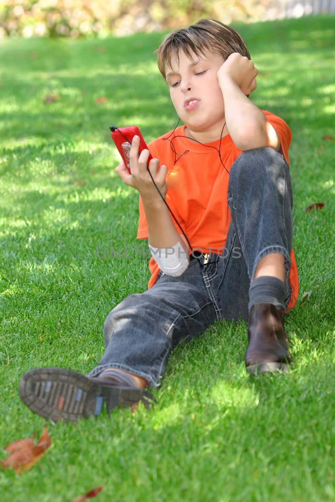 Young child relaxing in the park under the shade of a tree, head titled and looking at the mp3 player in his hand.