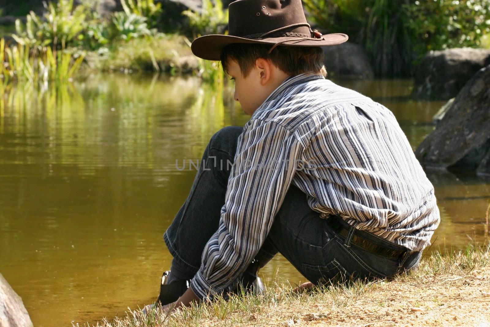 A child wearing jeans, shirt and hat sits quietly by the edge of a pond