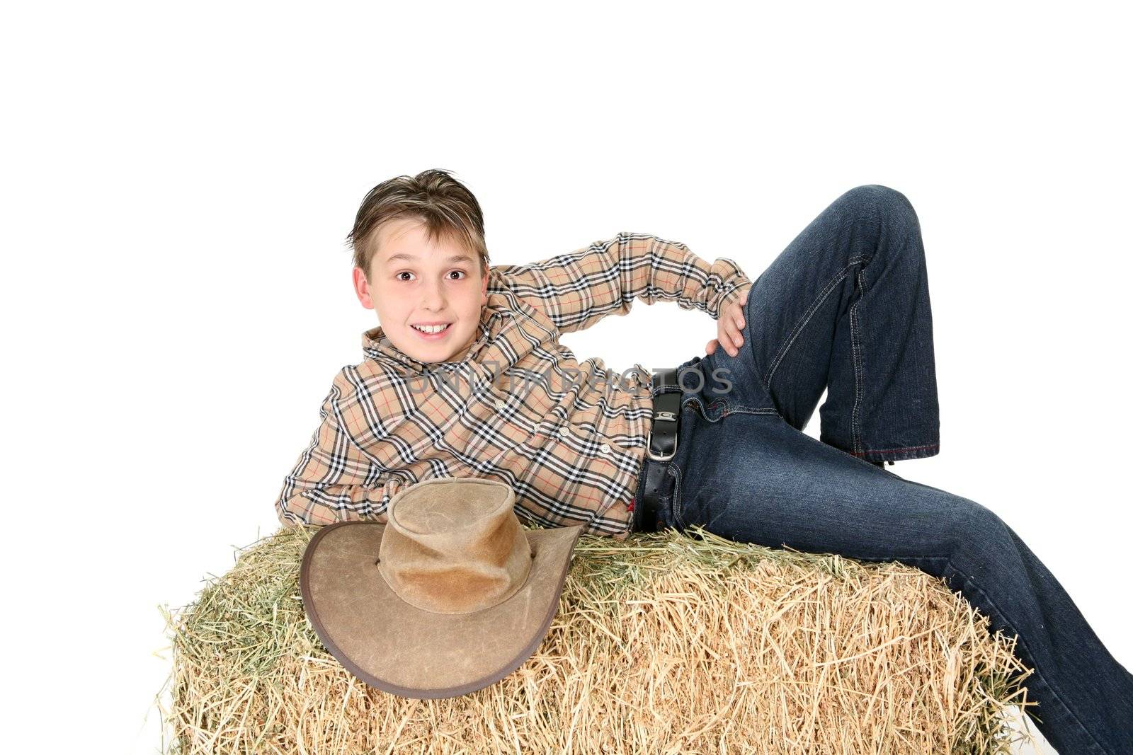 A boy from the country lays casually on a hay bale and smiles.  White background.