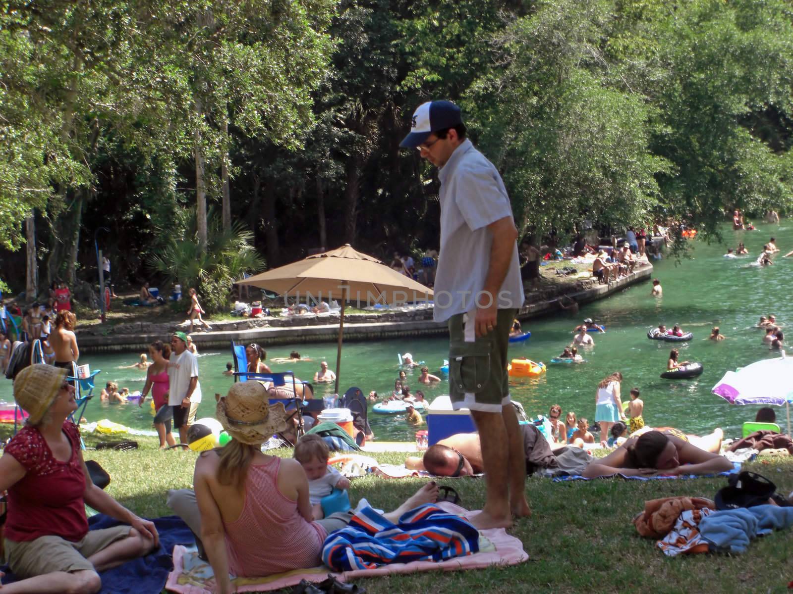 A crowd of people are lying in the sun while others are relaxing in the shade including some that are swimming inside a springwater fed pool at a state park