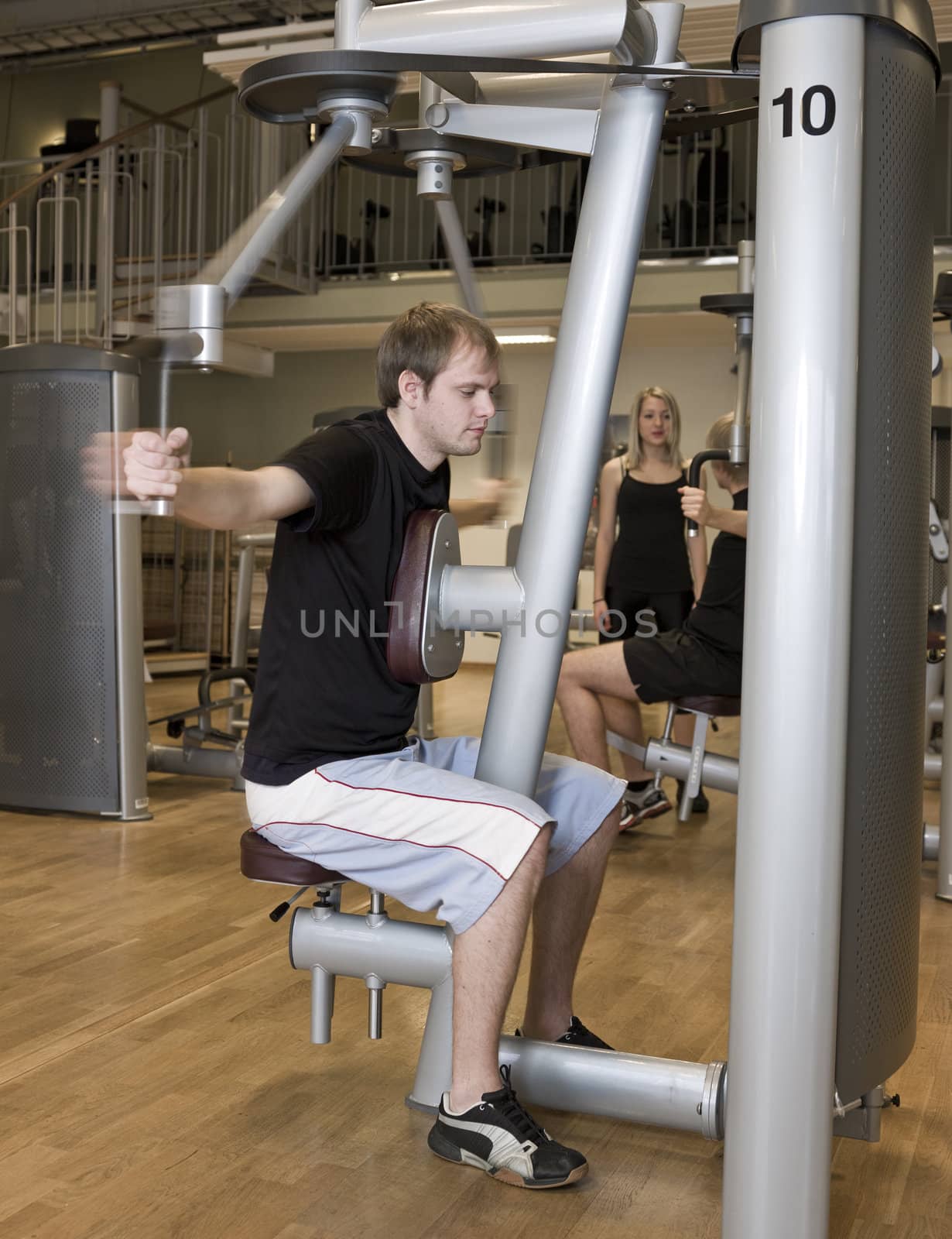 Young man using an exercise machine at a health club with a girl and a boy in the background