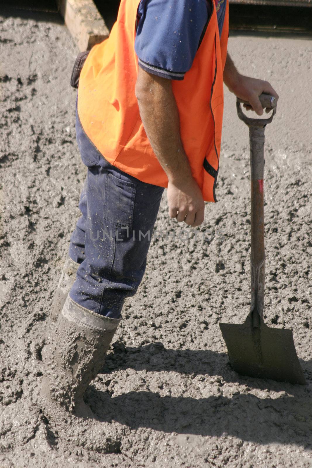 This construction worker stands in wet cement with knee high boots  covered in cement.  He is leaning on his his shovel
