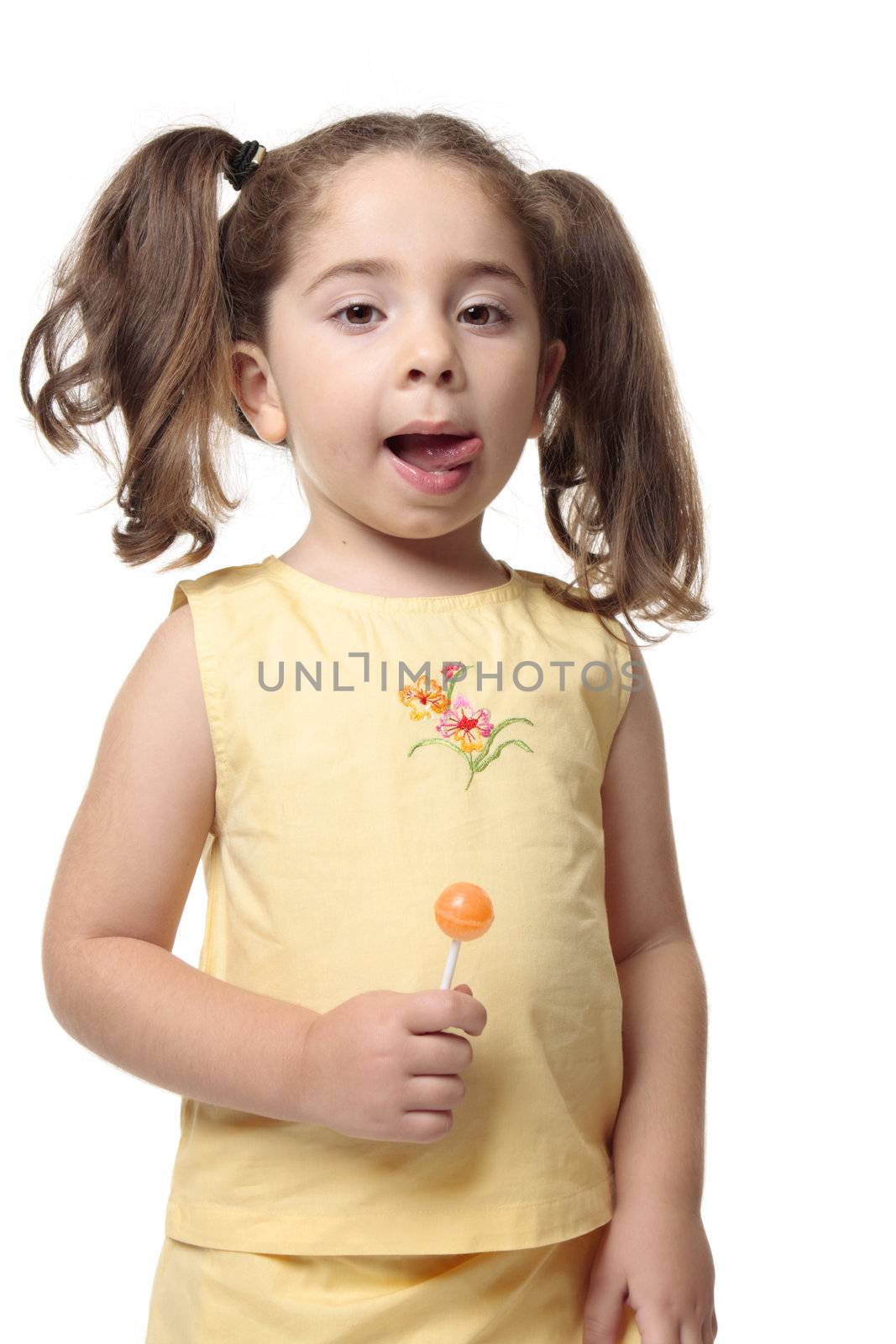 A little girl licks her lips.  She iis wearing a yellow outfit with hair in ponytails and she is holding a candy lollipop