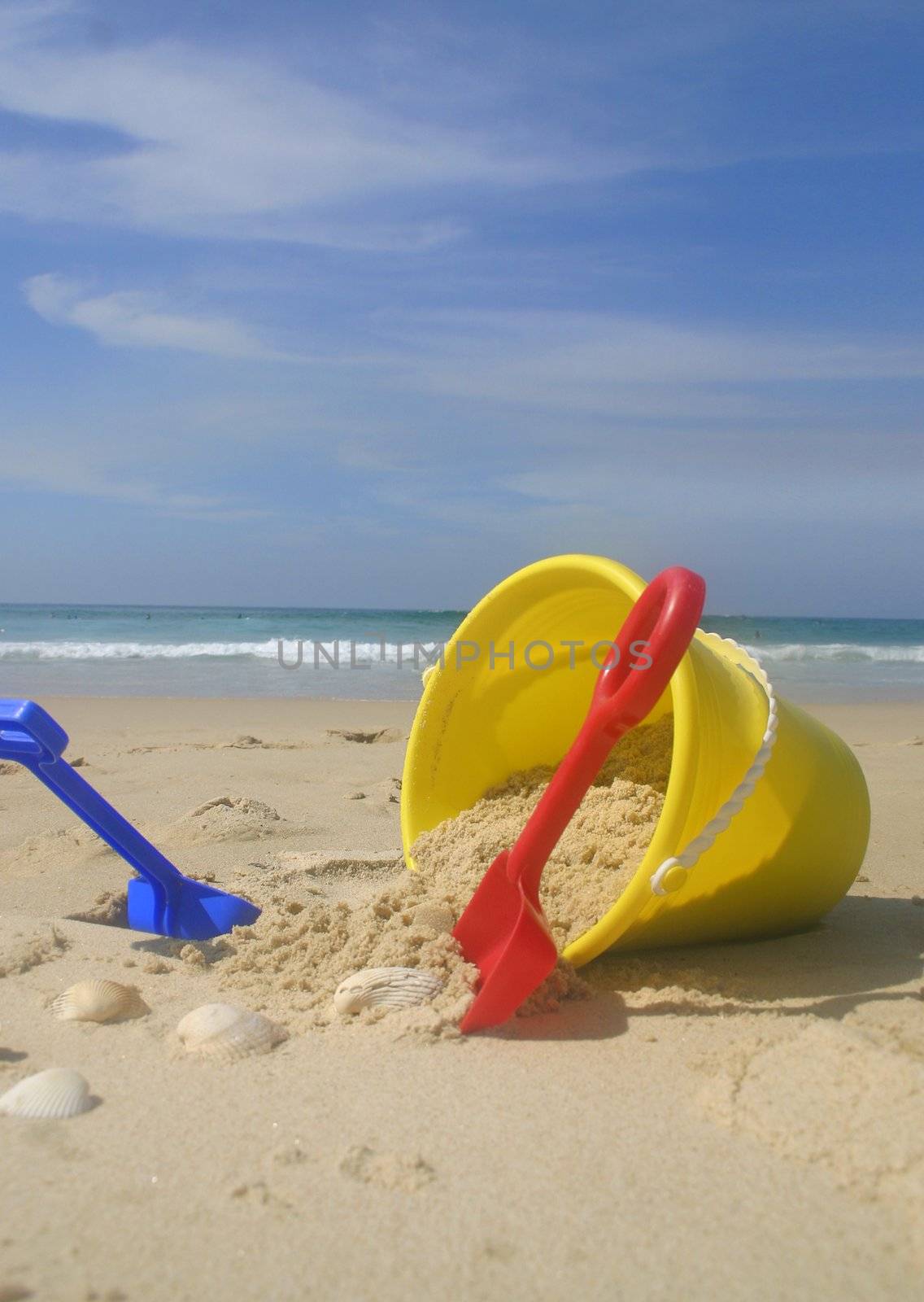 A childs yellow beach bucket and spades.    Focus is on the bucket of  sand and spades.   A couple of the shells and beach is oof range.