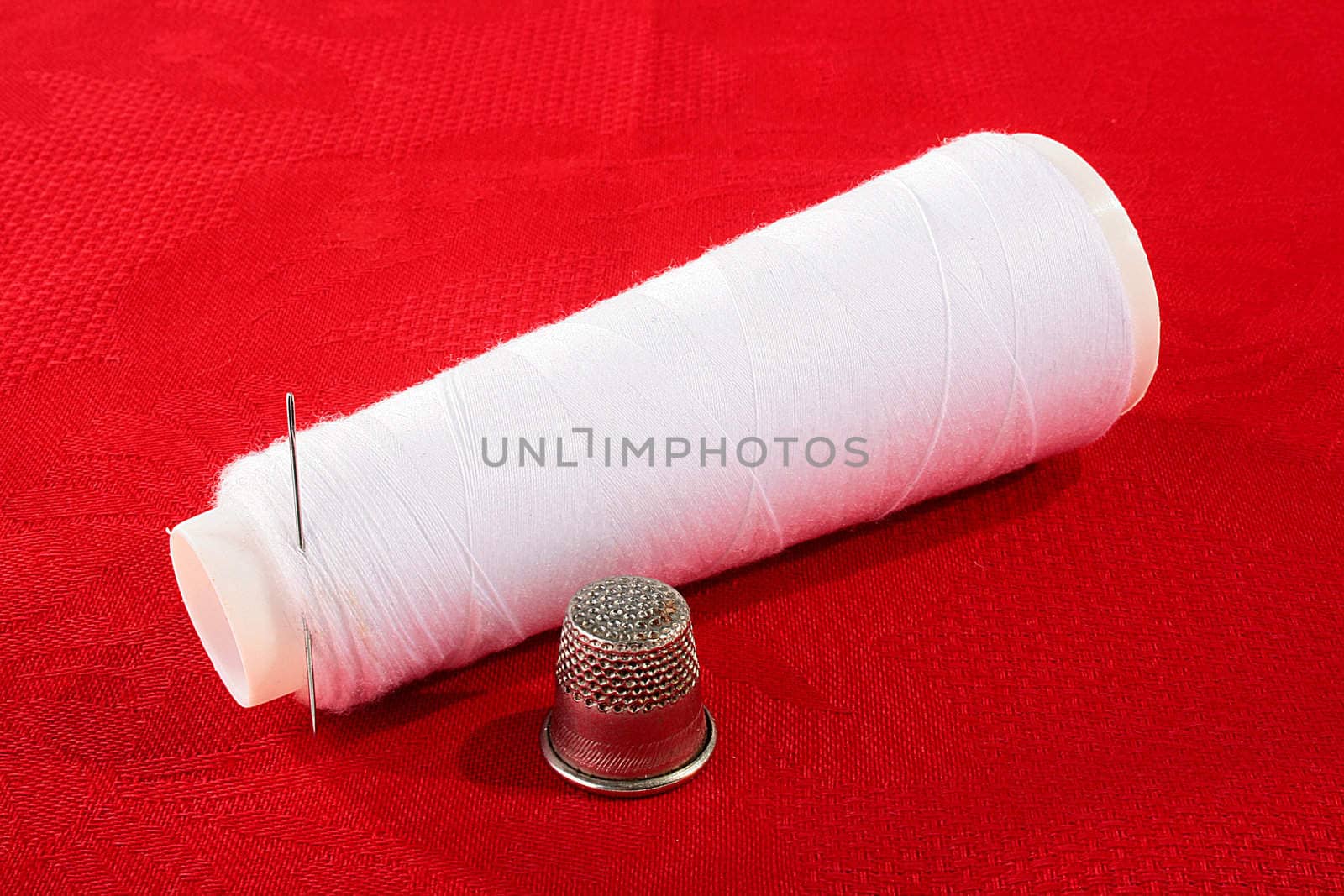 The coil with white threads and a needle on a red background, nearby a thimble for sewing.