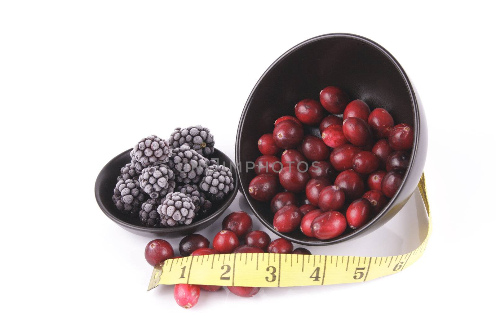 Red ripe cranberries spilling out of a small round black bowl on its side with a tape measure and frozen blackberries on a reflective white background